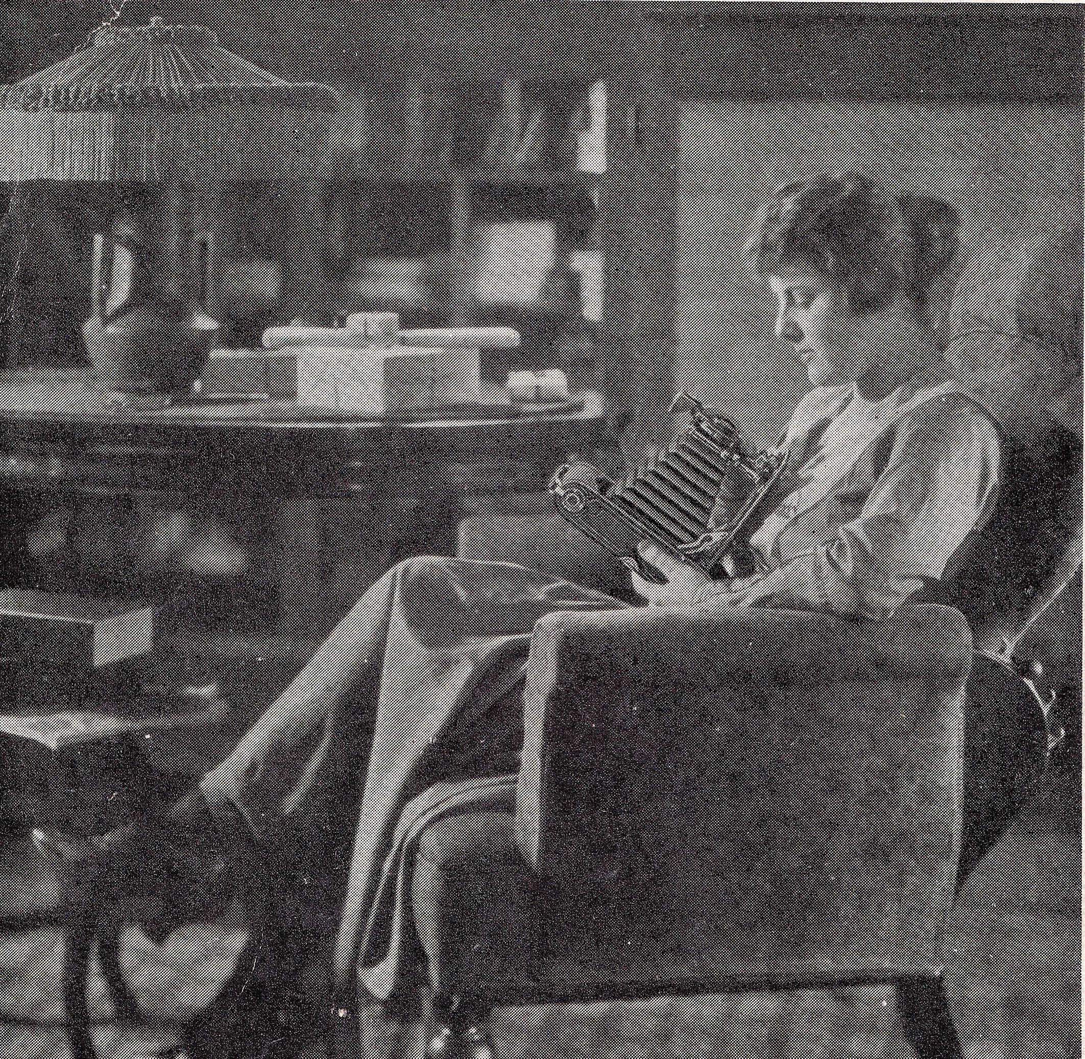 A young girl sitting in a rocking chair reading a book in a cozy room with bookshelves and kitchen items in the background.