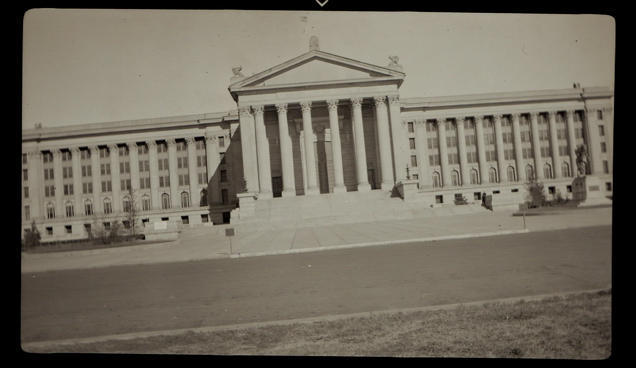 Black and white photograph of a large neoclassical government building with tall columns and steps leading up to the entrance, featuring sculptures on the roof.