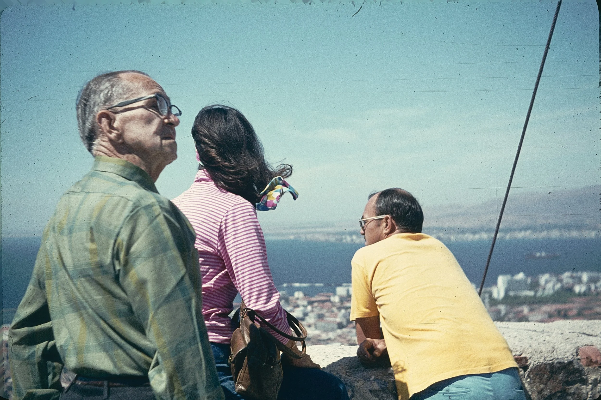Four people sitting outdoors on a sunny day, with a cityscape and water in the background. One man in glasses and a striped shirt, a woman with dark hair and a colorful scarf, another man with glasses, and a person in a yellow shirt.