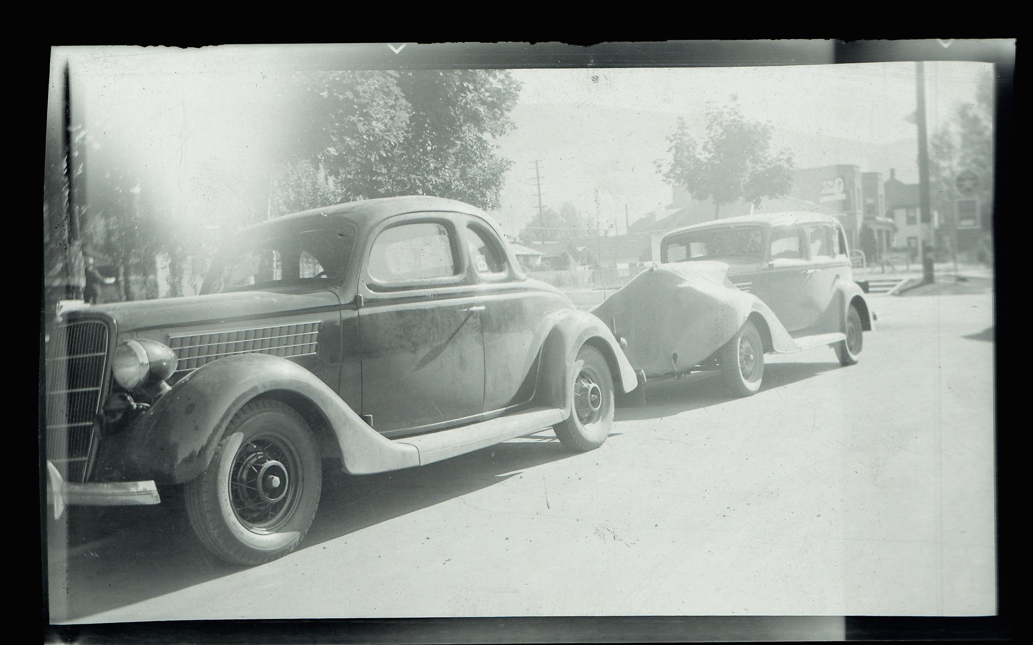 Black and white photo of vintage cars parked on a street, with trees and buildings in the background.