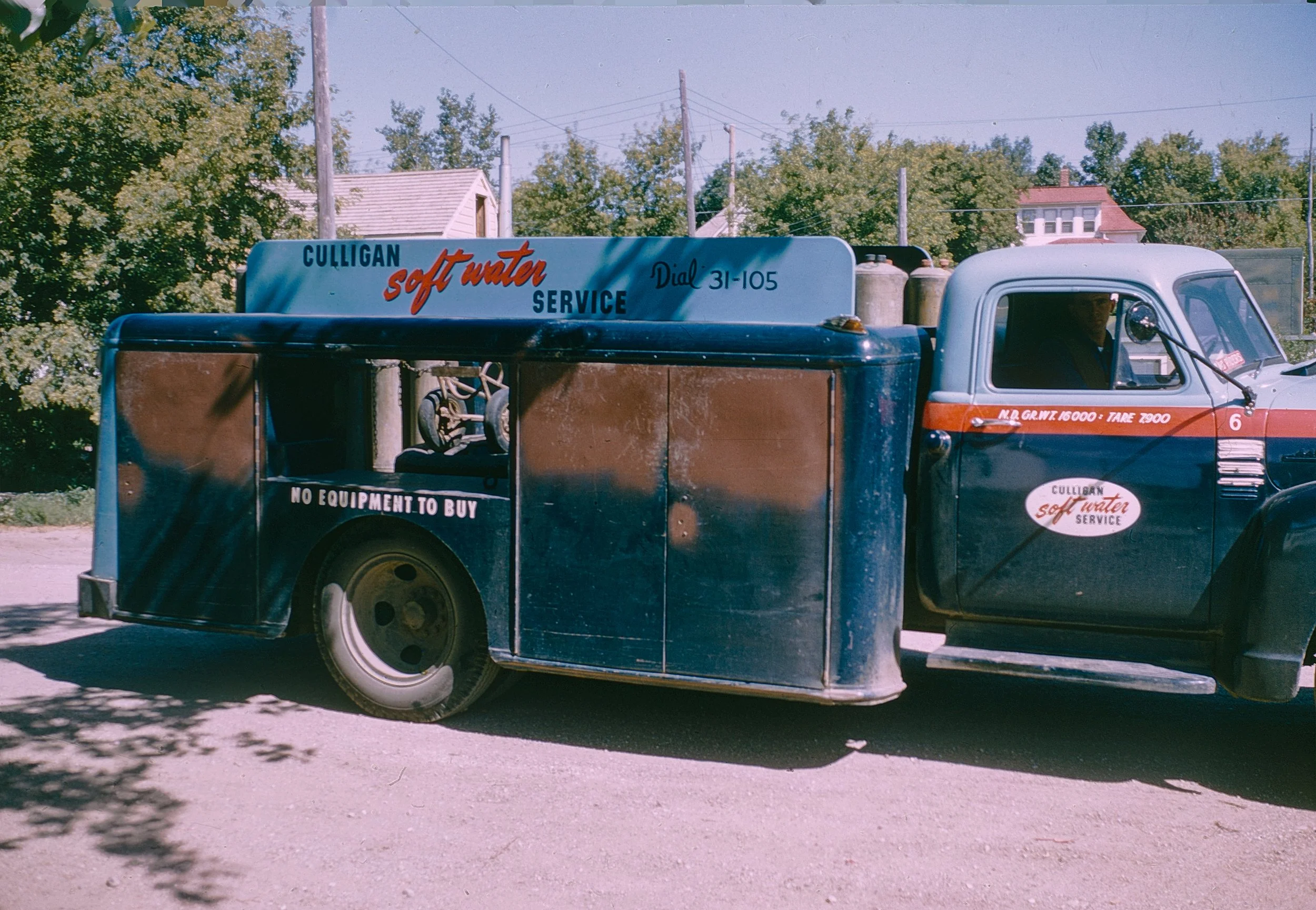 A vintage delivery truck with a blue and orange color scheme parked on the side of a street. The truck advertises Culligan soft water service, with a sign on top and an oval logo on the door. The truck has two cylinders mounted on the back, and the b