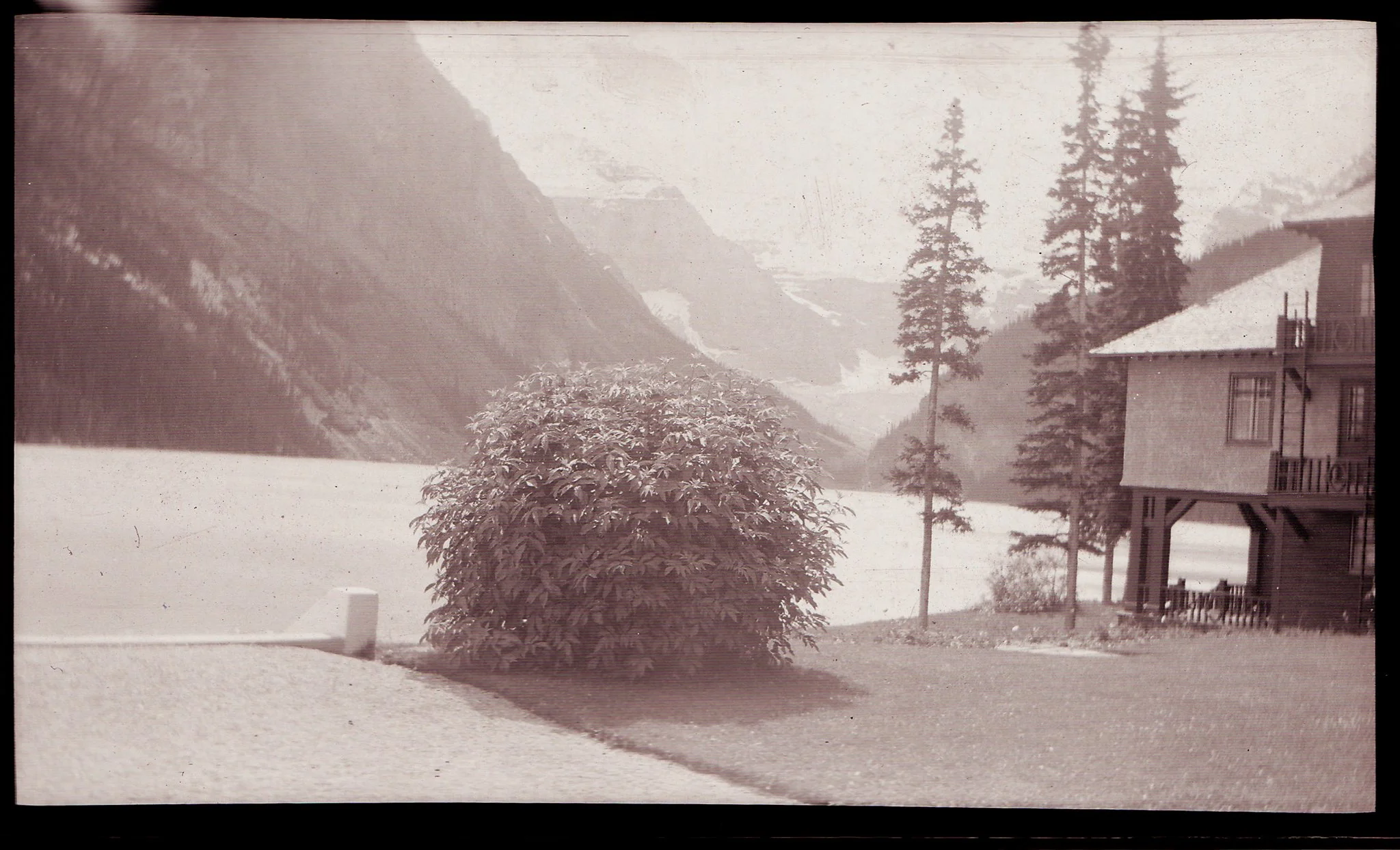 A mountain landscape with snow-capped peaks, evergreen trees, a bush, a house, and a body of water in the foreground.