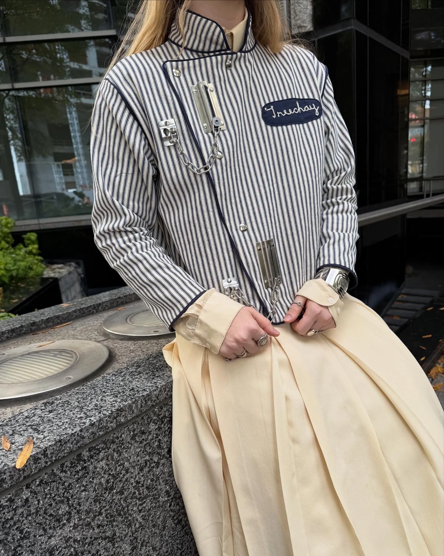 A person with long blonde hair dressed in a vintage-style uniform, including a striped jacket with metal chains and patches, and a light yellow apron/skirt, standing outdoors beside a granite countertop with built-in lights.