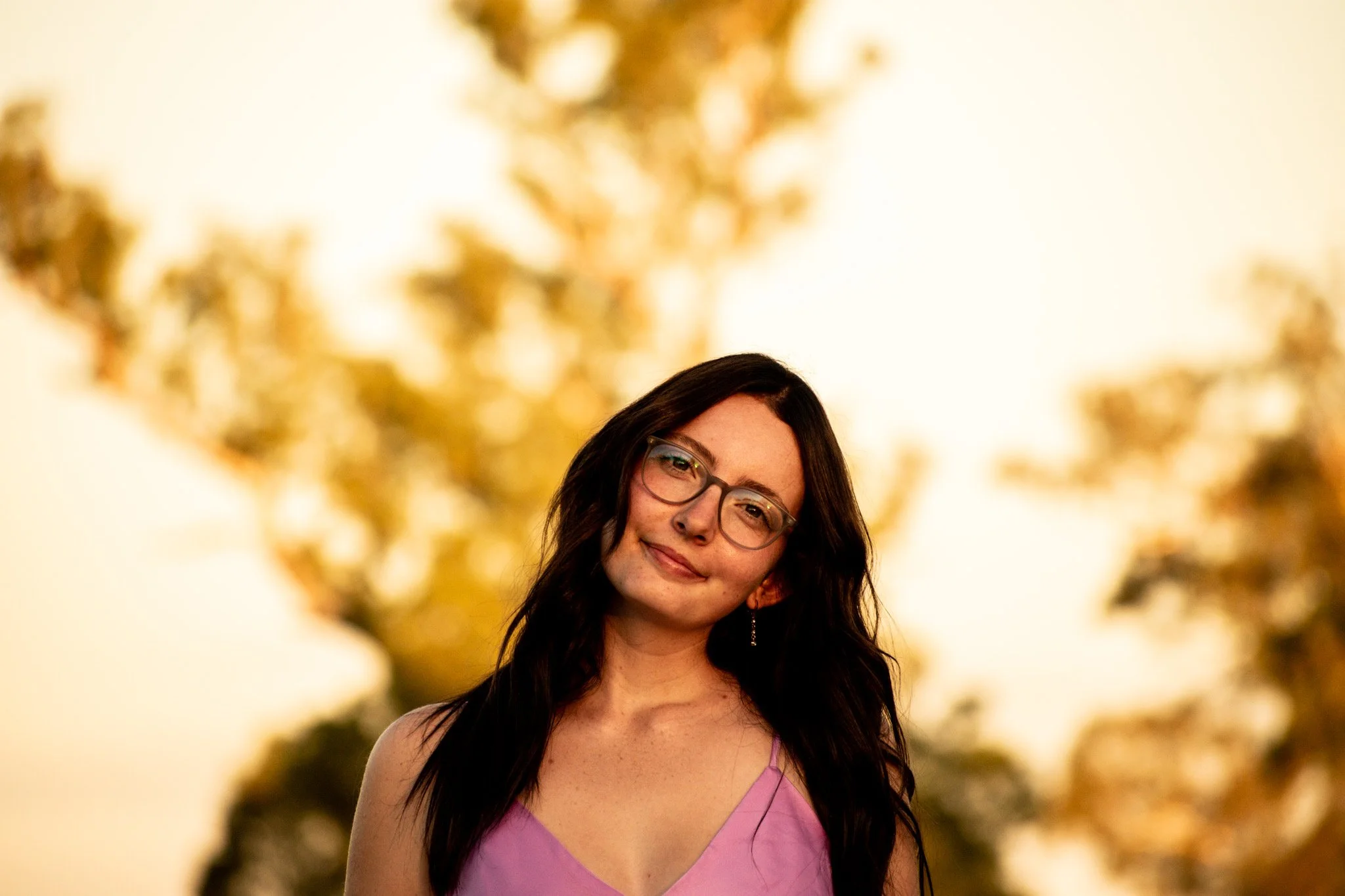 A young woman with long black hair, glasses, and earrings standing outdoors during sunset, wearing a pink sleeveless top, with blurred trees in the background.