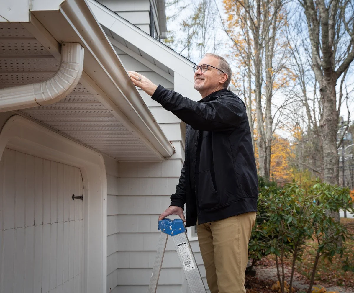 A man stands on a stepladder, inspecting the gutter system of a house outside during autumn, with trees in the background.