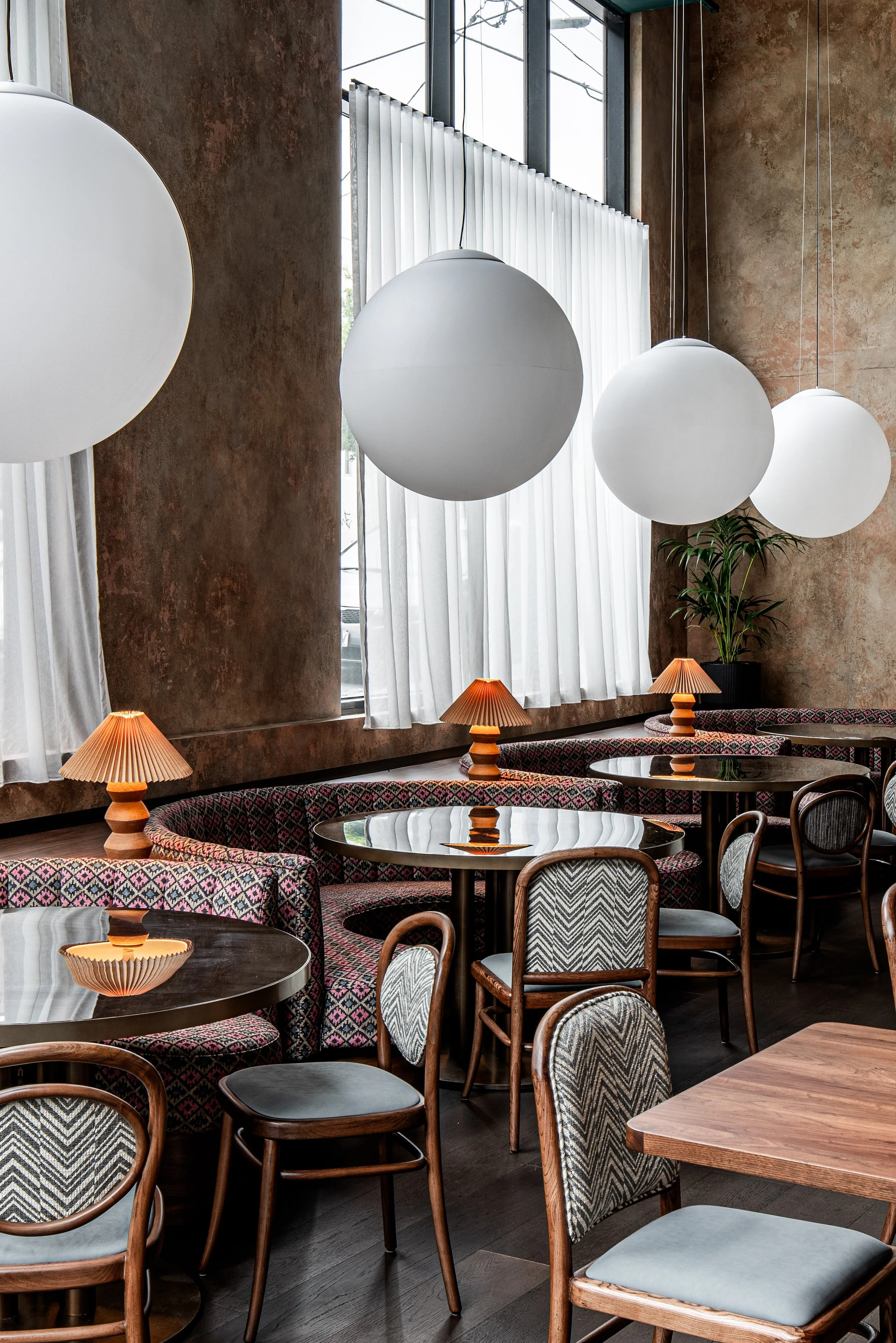Interior of a modern restaurant or cafe with round tables, upholstered chairs, and curved booth seating. Large spherical pendant lights hang from the ceiling, and small table lamps add warm lighting. Tall windows with white curtains let in natural light, and a potted plant is in the corner.