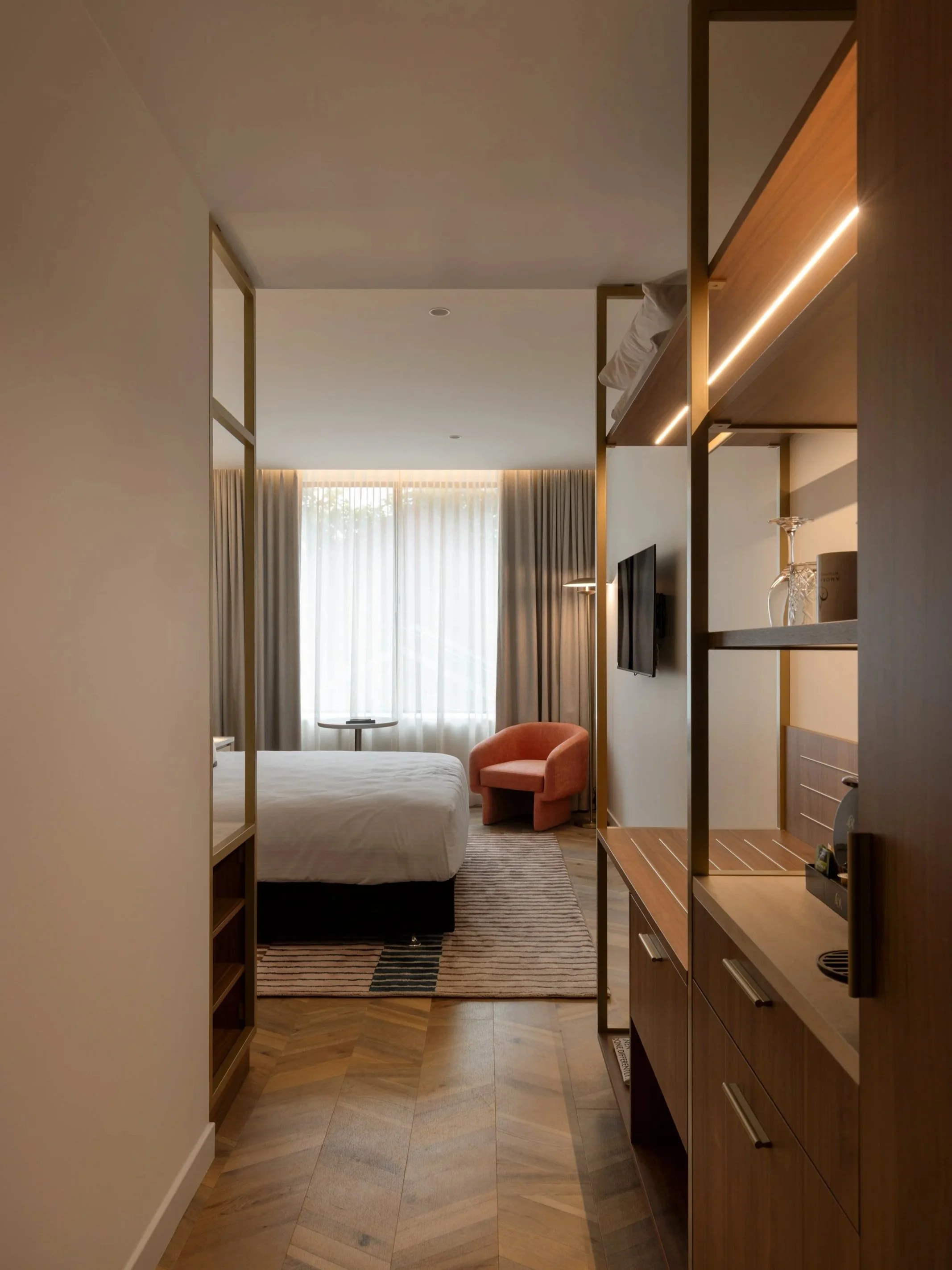 Interior view of a hotel room with a bed, window with curtains, a pink armchair, a side table, and a wooden shelving unit.