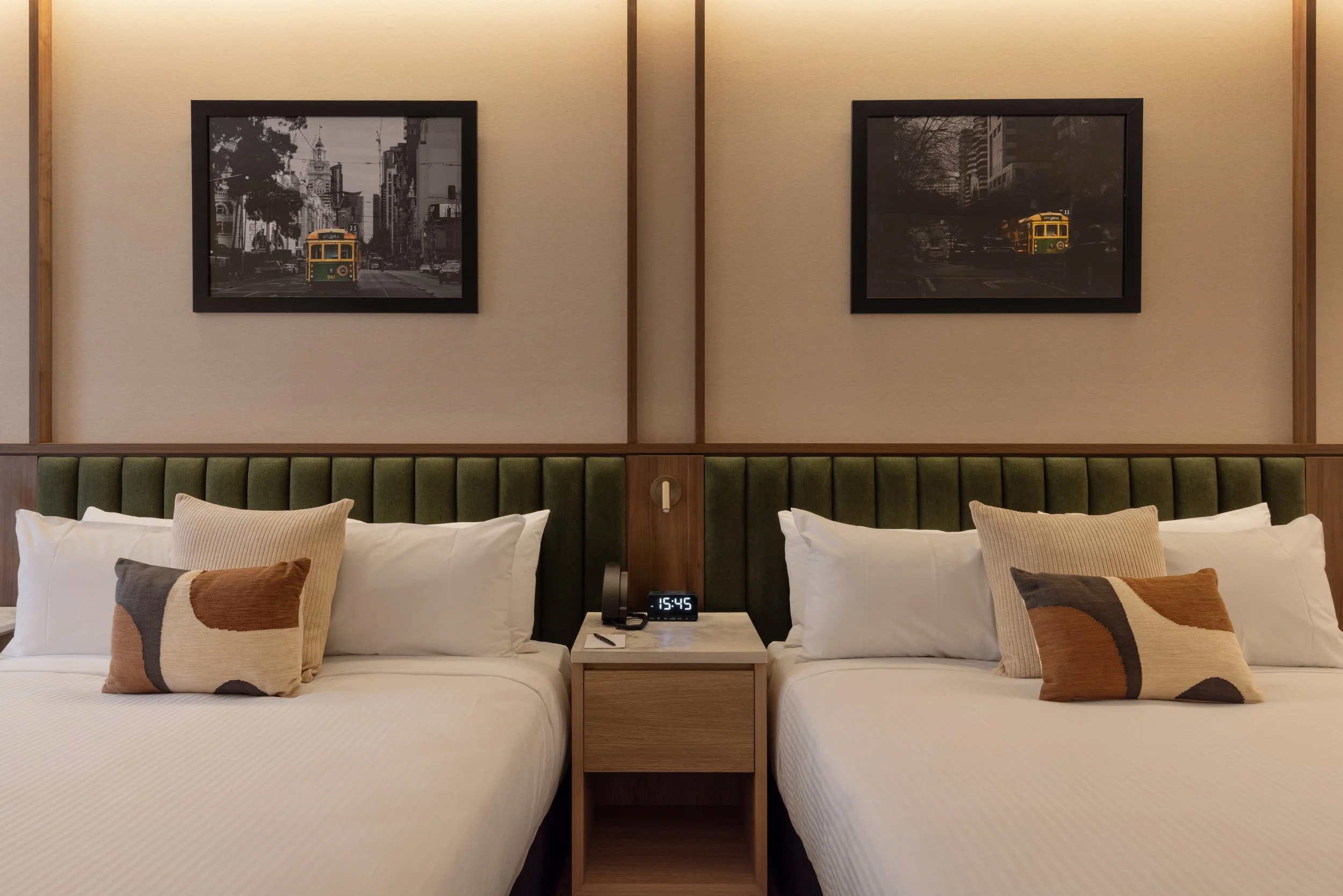 Two neatly made beds with white linens and decorative pillows in a hotel room. A wooden nightstand with a clock, phone, and glasses is between the beds. Two framed black and white cityscape photographs hang above the beds.