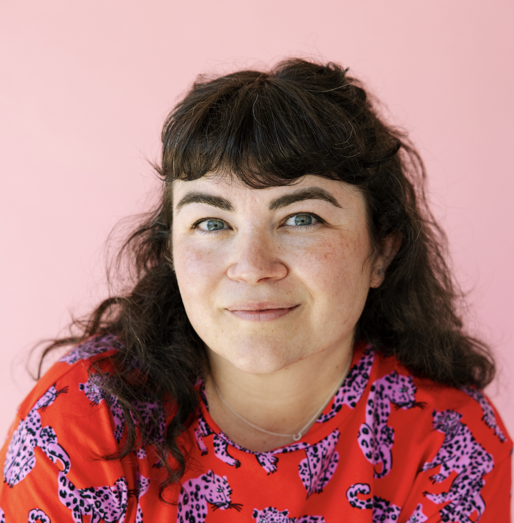portrait of a woman in her early 40's with bangs and a red tee with pink leopards on it in front of a pink backdrop