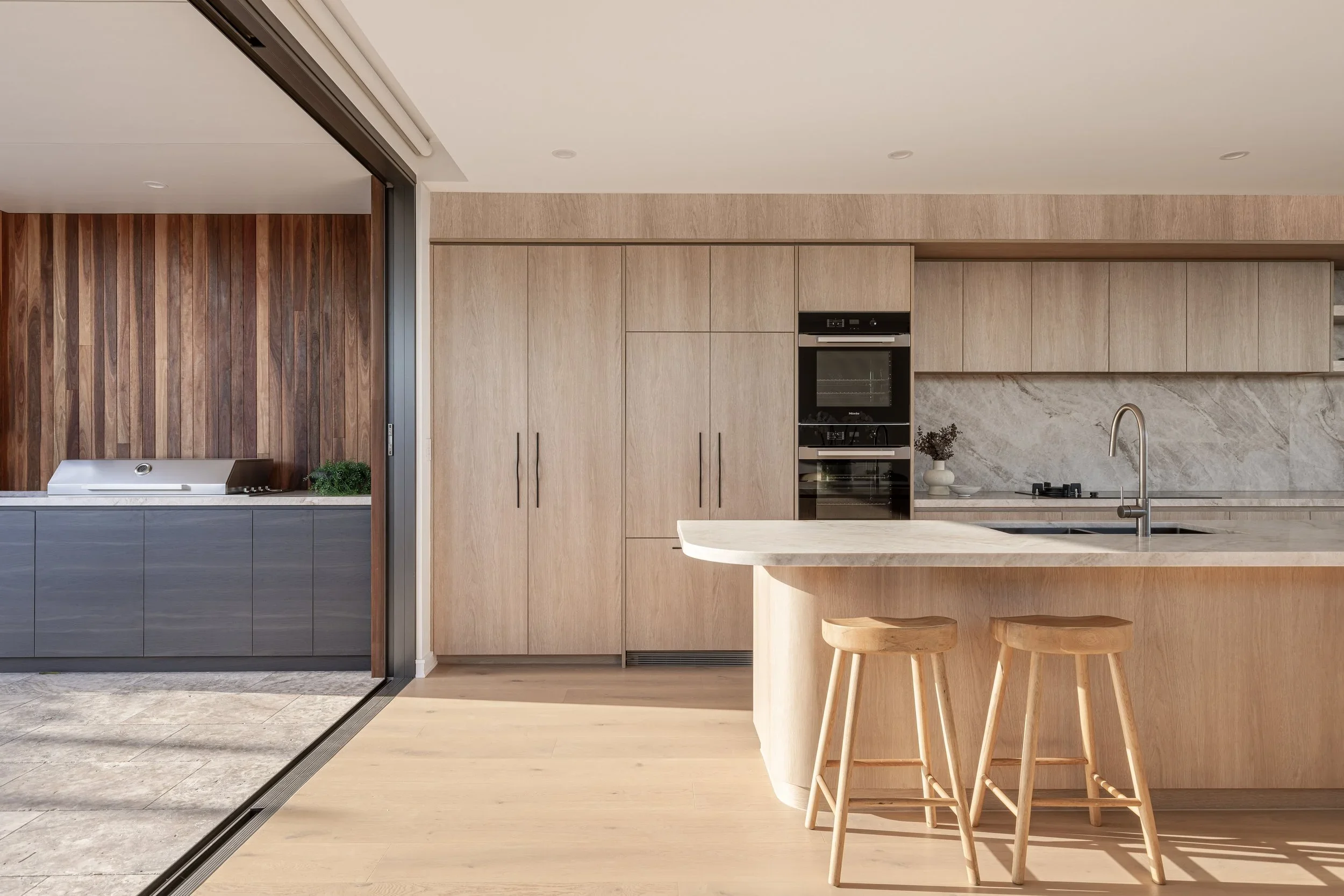 Modern kitchen with light wooden cabinets, marble countertop, and two wooden bar stools.