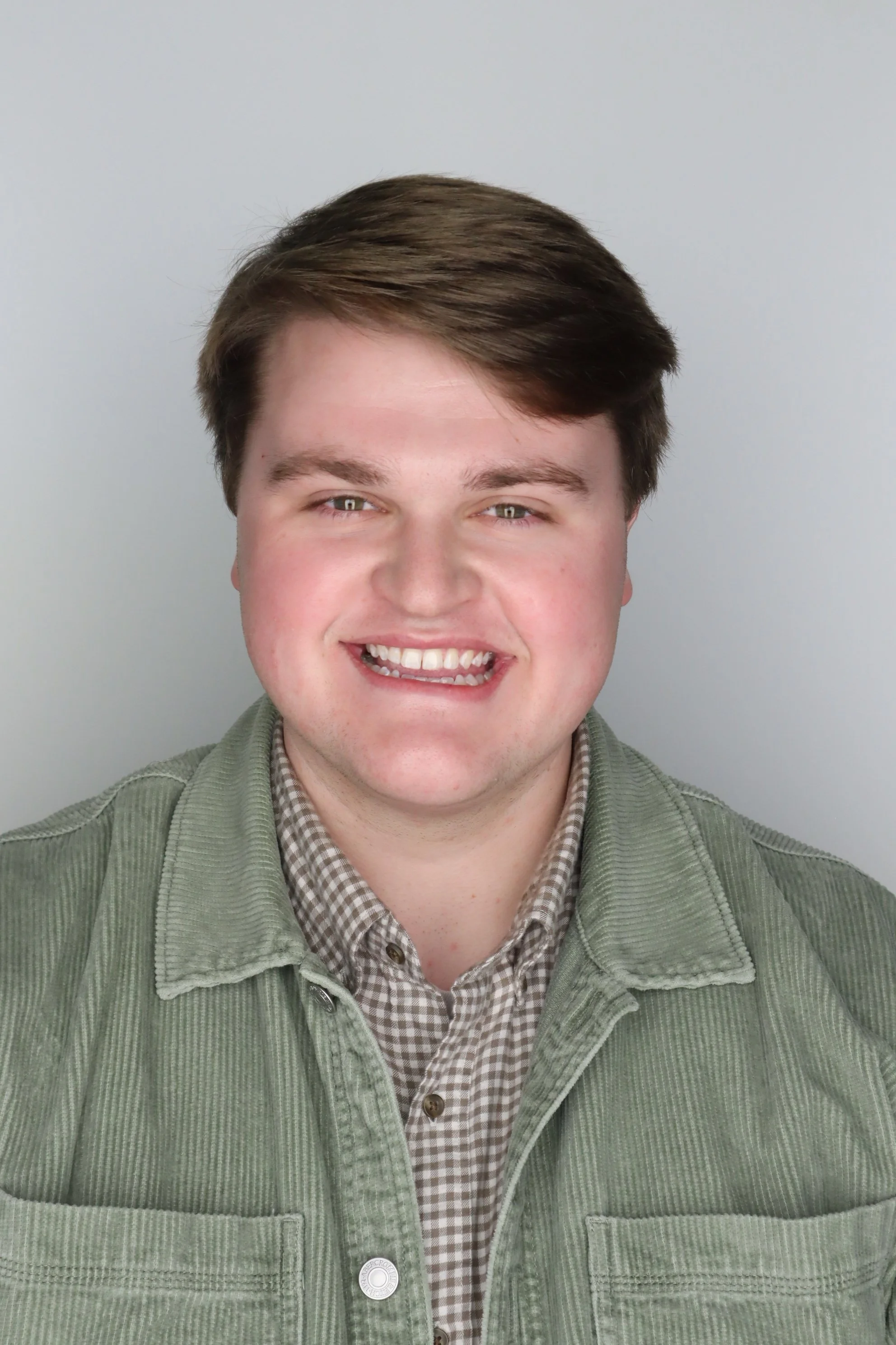 A young man with brown hair, smiling, wearing a green corduroy jacket and a checkered shirt, standing against a plain light background.