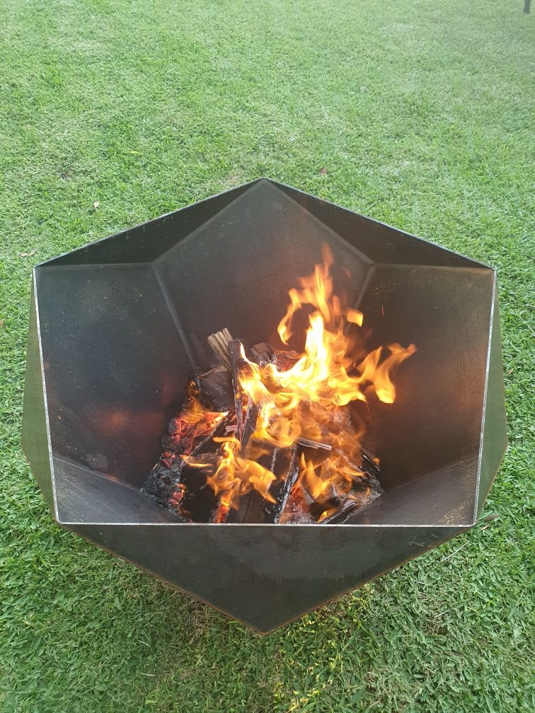 A metal fire pit with burning wood and flames, placed on green grass outdoors.