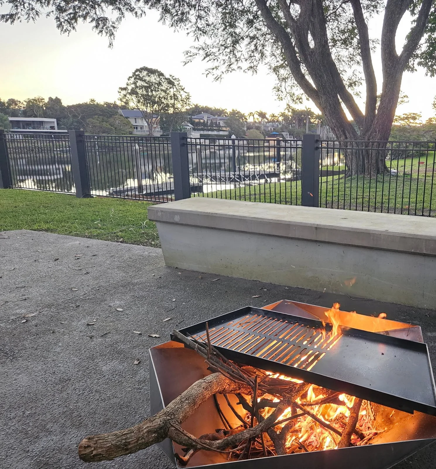 A backyard with a fire pit containing burning wood and small flames on a concrete surface. A concrete bench is beside the fire pit. In the background, there is a black metal fence, green grass, a large tree, and houses across a waterway.