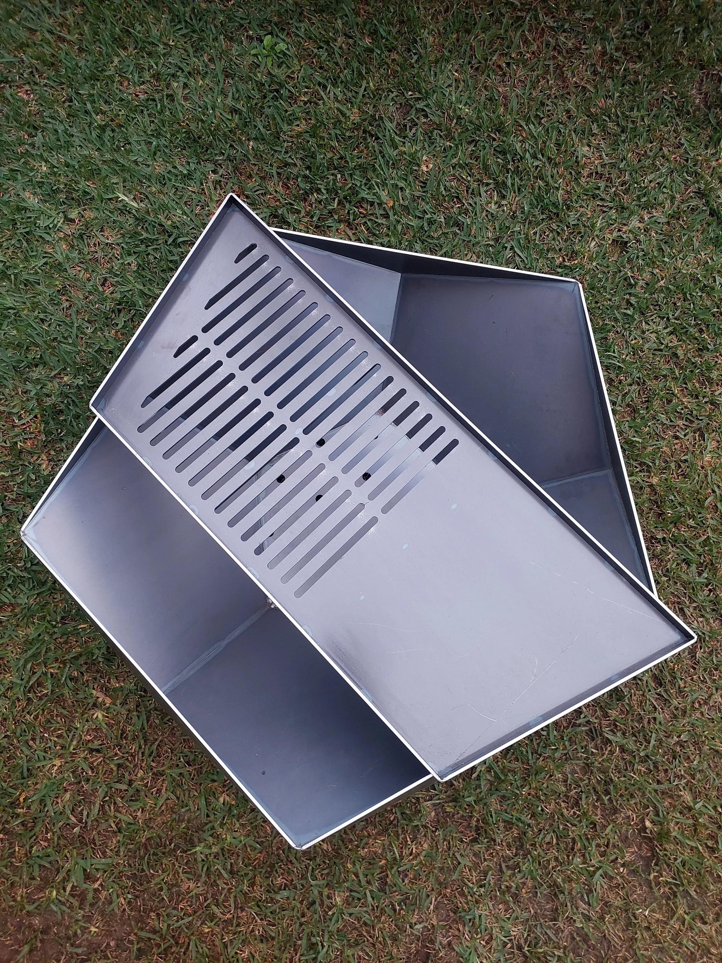Metal grilling barbecue with a large flat surface and a slotted grilling rack, placed on grass.