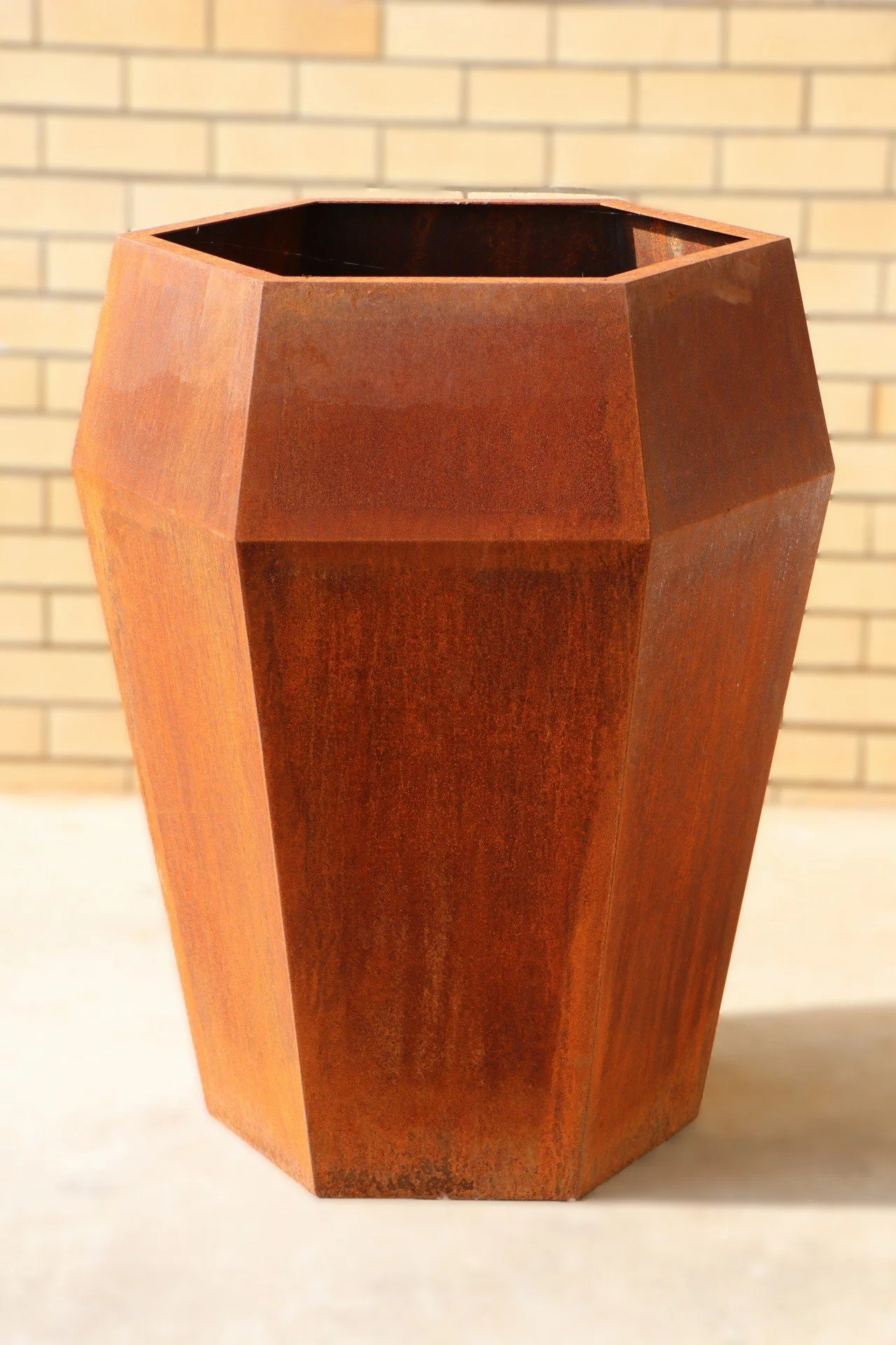 A tall, rust-colored, faceted metal planter with a geometric design, placed on a light surface with a brick wall in the background.