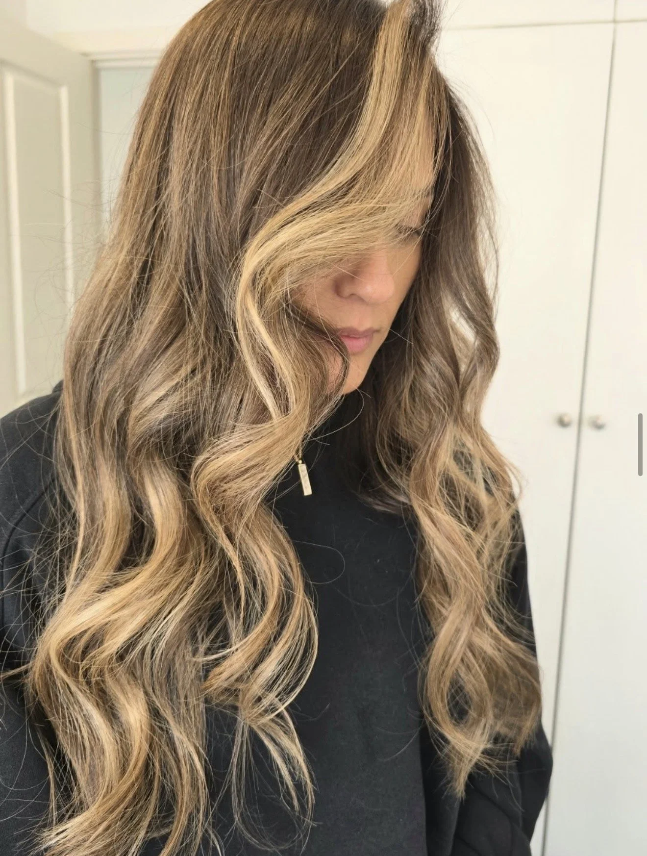 A woman with long, wavy, blonde hair with darker roots, wearing a black top, standing indoors against a white door.