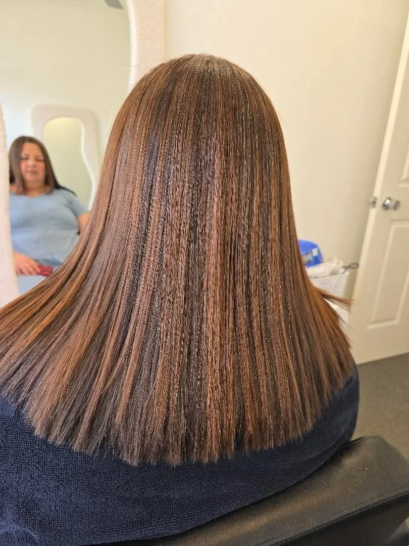 Back of a woman with straight, shiny, brown hair sitting in a salon chair, facing a mirror with her reflection visible.