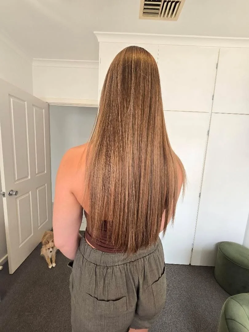 Back view of a woman with long, straight, light brown hair, wearing a sleeveless top and gray shorts, standing in a room with a small dog in the background.