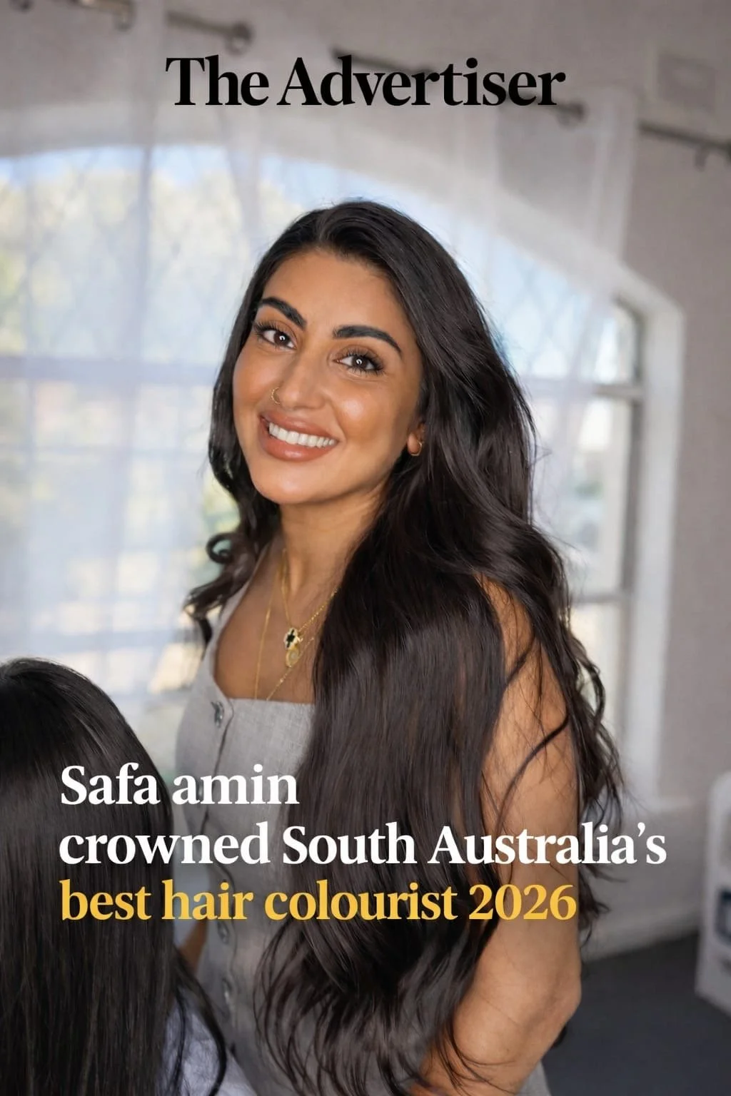 A woman with long dark hair, smiling, wearing a gray sleeveless top and jewelry, and two children with dark hair are in the foreground, indoors with a bright window in the background.