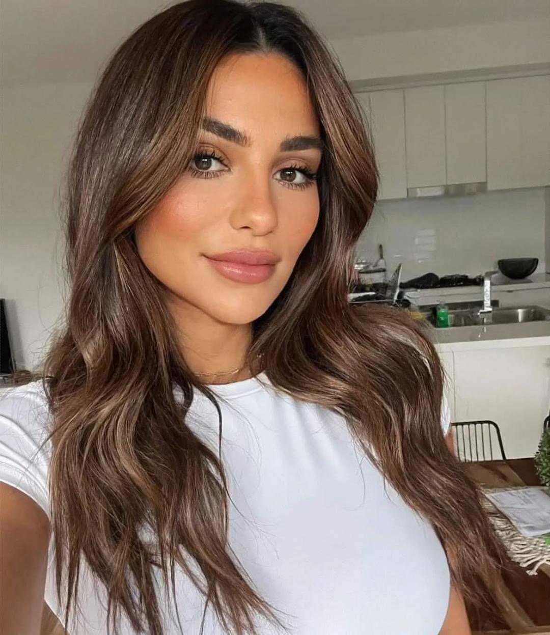 A young woman with long, wavy brown hair and light makeup is taking a selfie in a modern kitchen, wearing a white t-shirt.