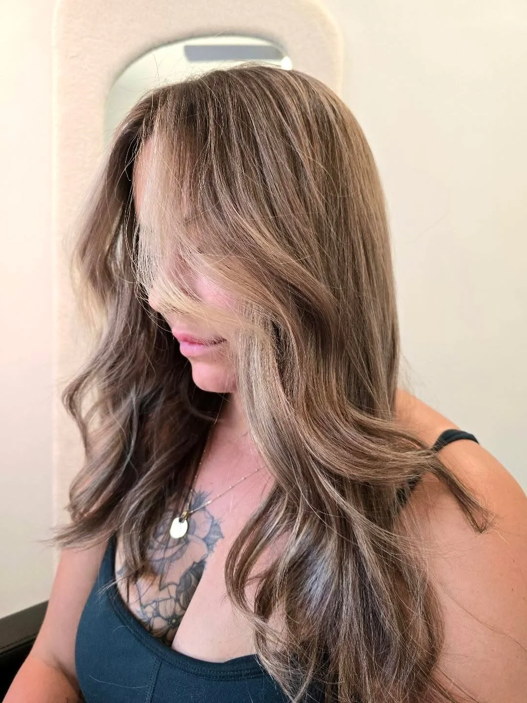 A woman with wavy, light brown hair with blonde highlights, looking down, wearing a black tank top and a silver necklace with a pendant, with a tattoo of flowers on her chest.