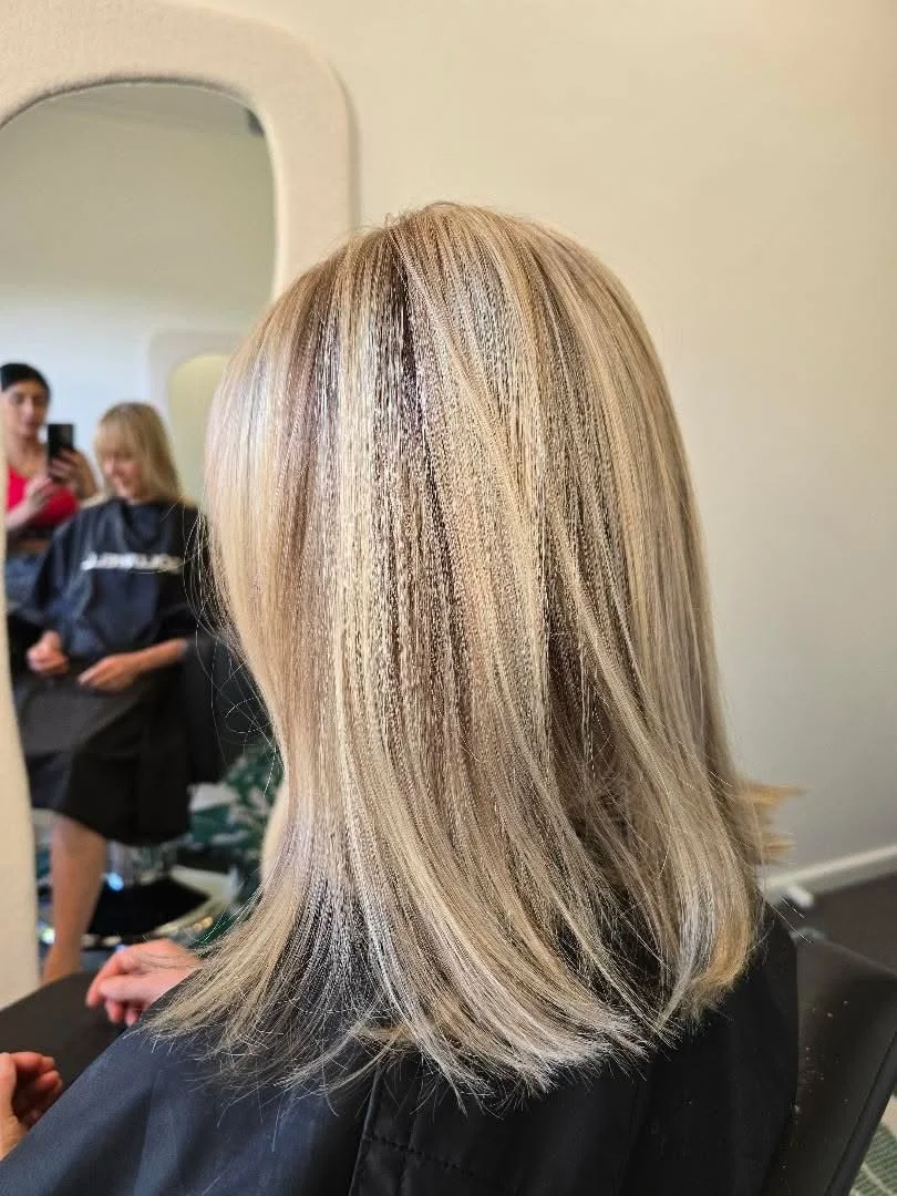 Close-up of a woman's blonde, straight hair with subtle highlights, in a salon setting with a large mirror and two people in the background.