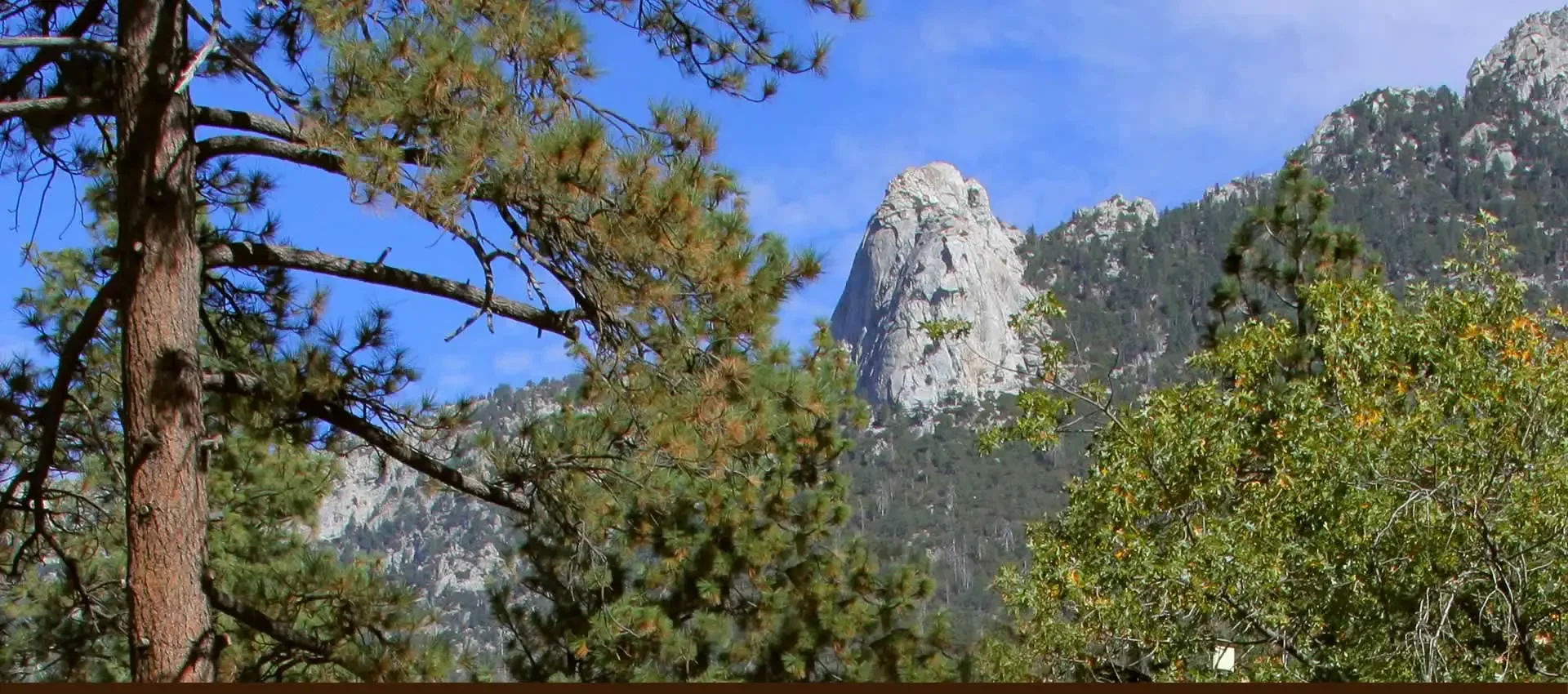Mountain landscape with large rock formation, pine trees, and blue sky.