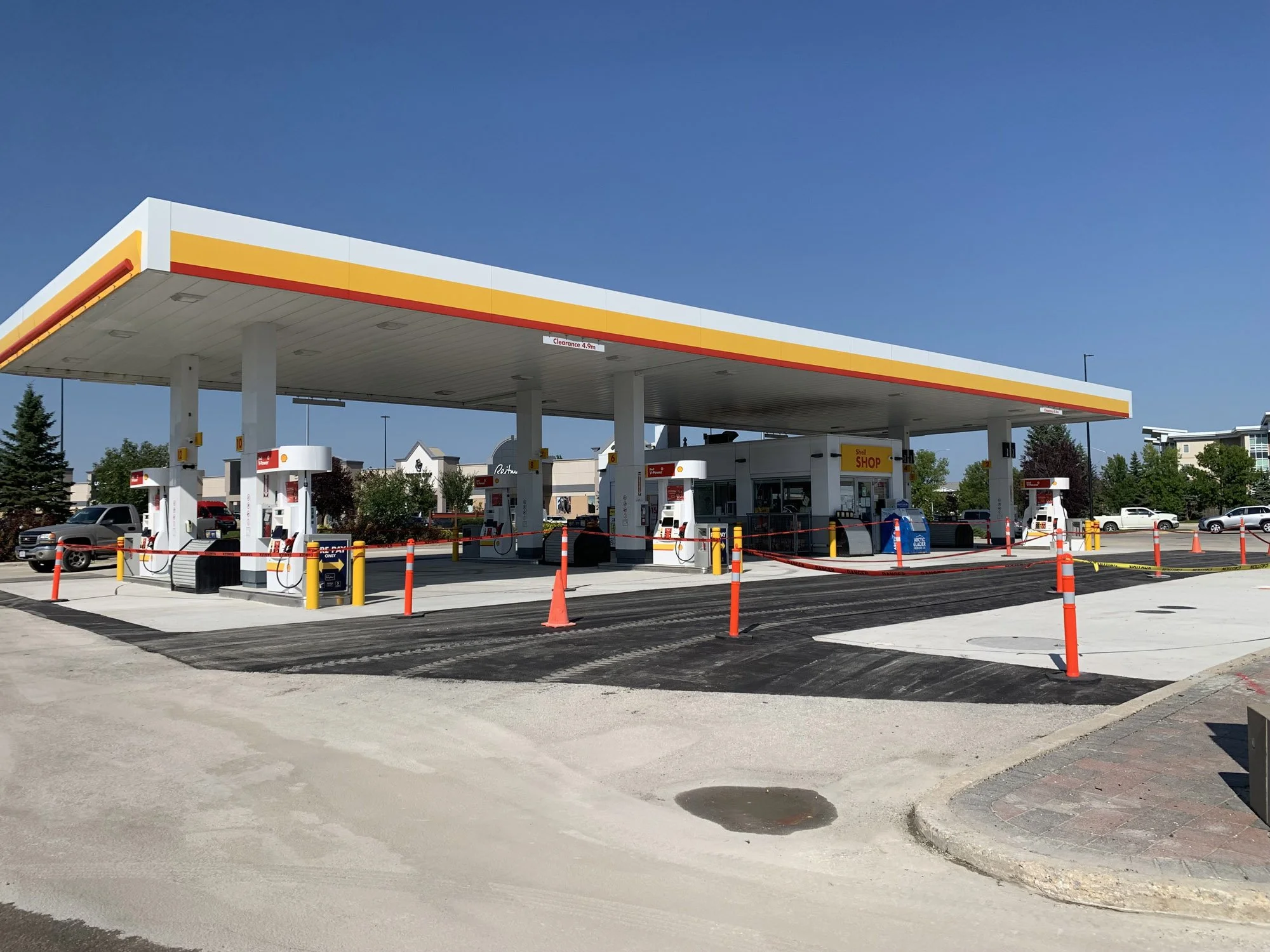 A Shell gas station with four fuel pumps under a canopy, surrounded by orange traffic cones and caution tape, with parked cars and trees in the background under a clear blue sky.