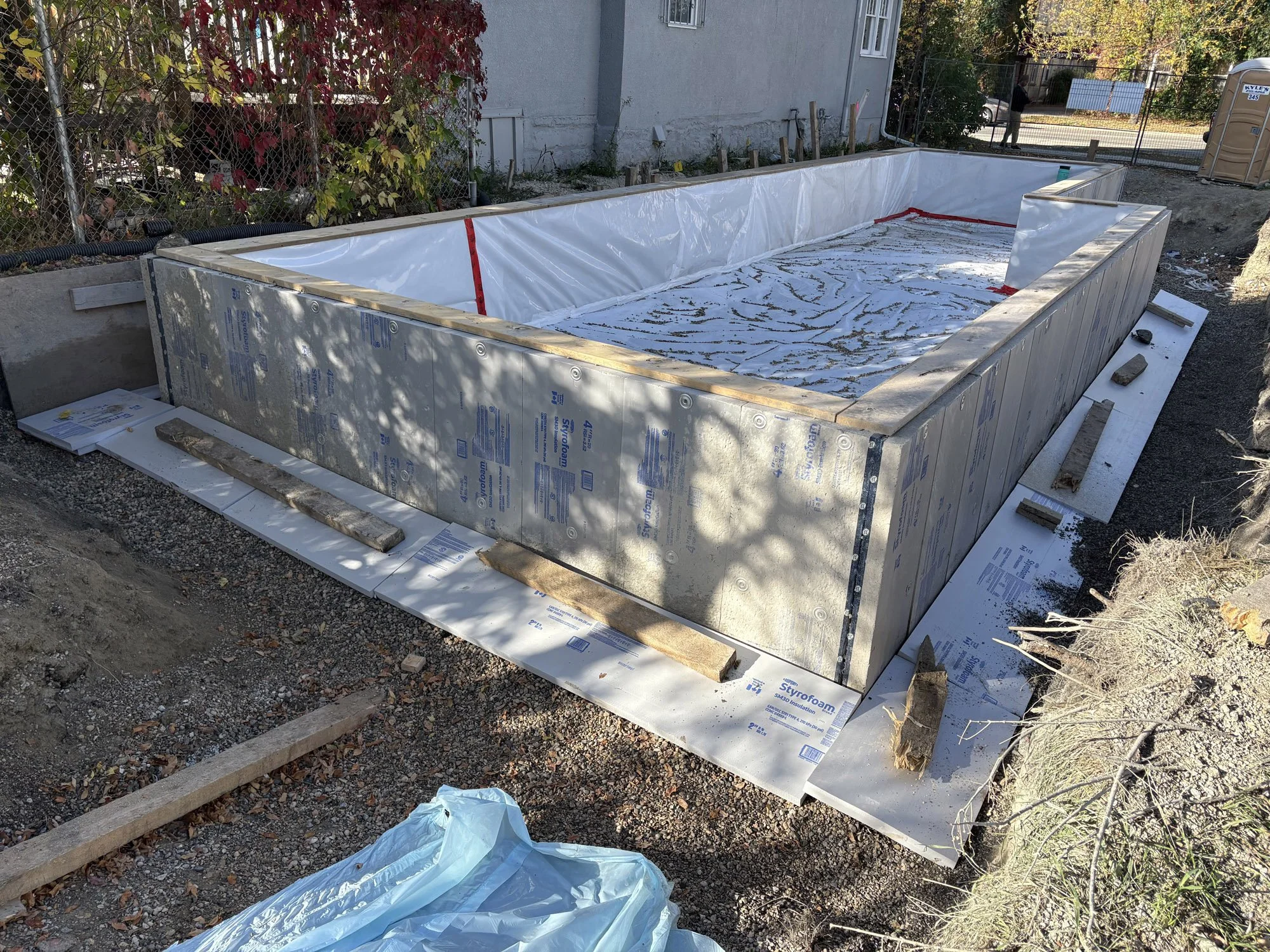 Construction site of an in-ground swimming pool with partial walls and lining, surrounded by construction materials, gravel, and dirt, near a house, with trees and a portable toilet in the background.