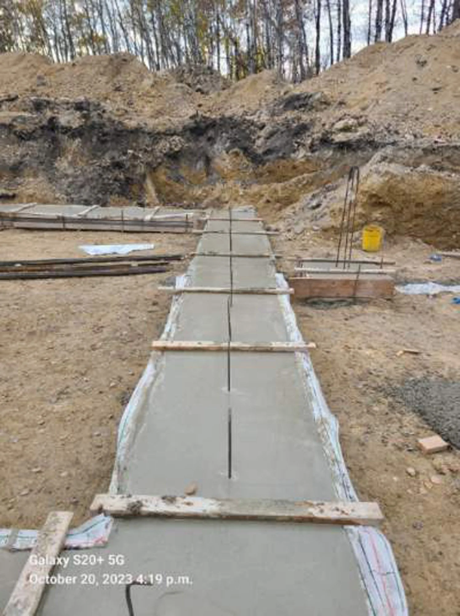 Construction site with freshly poured concrete pathway, wooden supports, and exposed soil and rocks, with trees in the background.