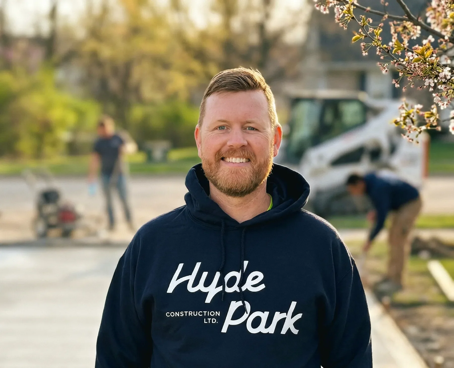 A smiling man with a beard wearing a navy hoodie with the words 'Hyde Park' and 'Construction Ltd.' printed on it, standing outdoors during daytime with a blurred background of people working and construction equipment.