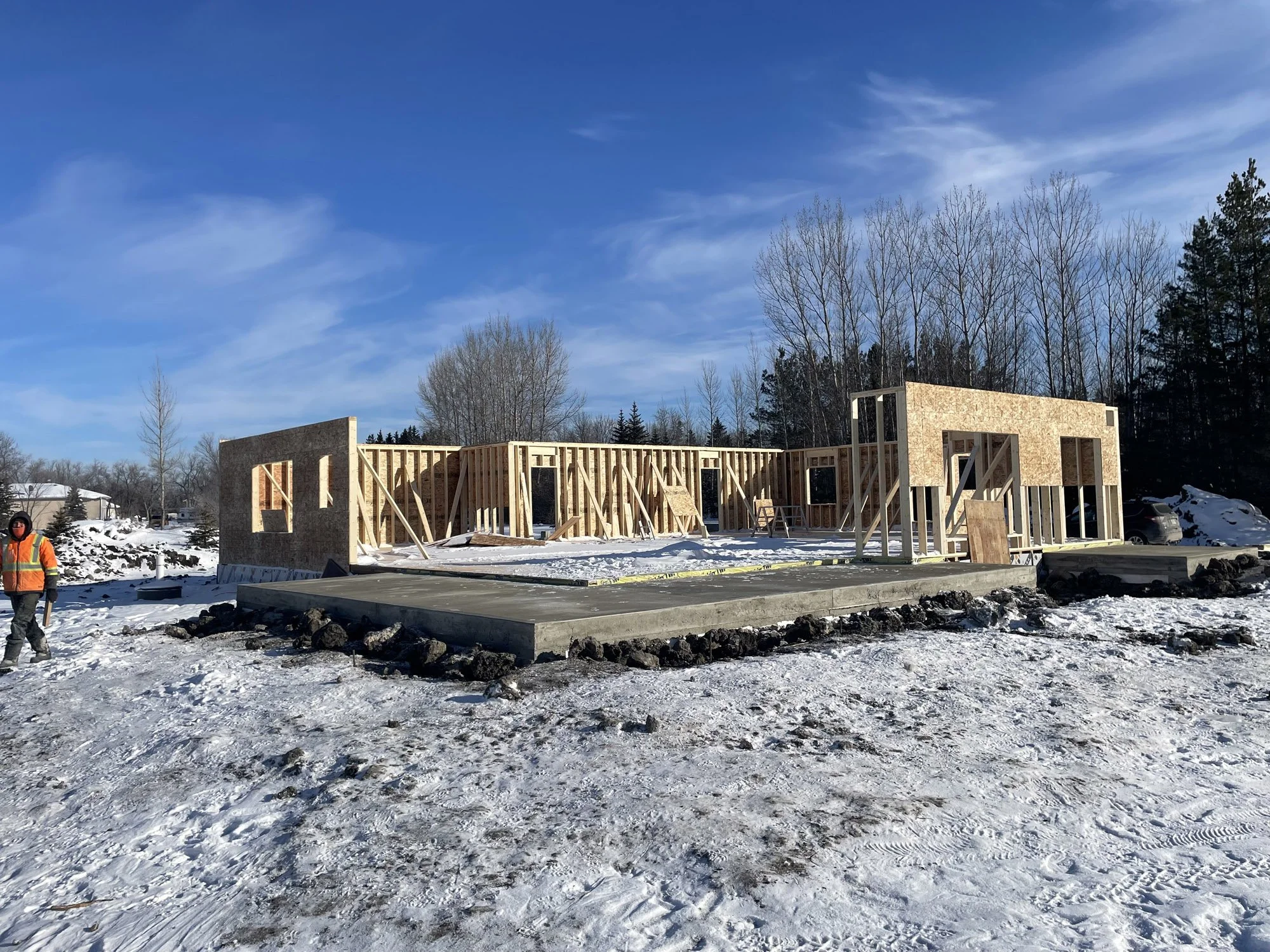 Frame of a house under construction with wood framing, surrounded by snow, a worker in an orange safety vest walking nearby, and trees and a blue sky in the background.