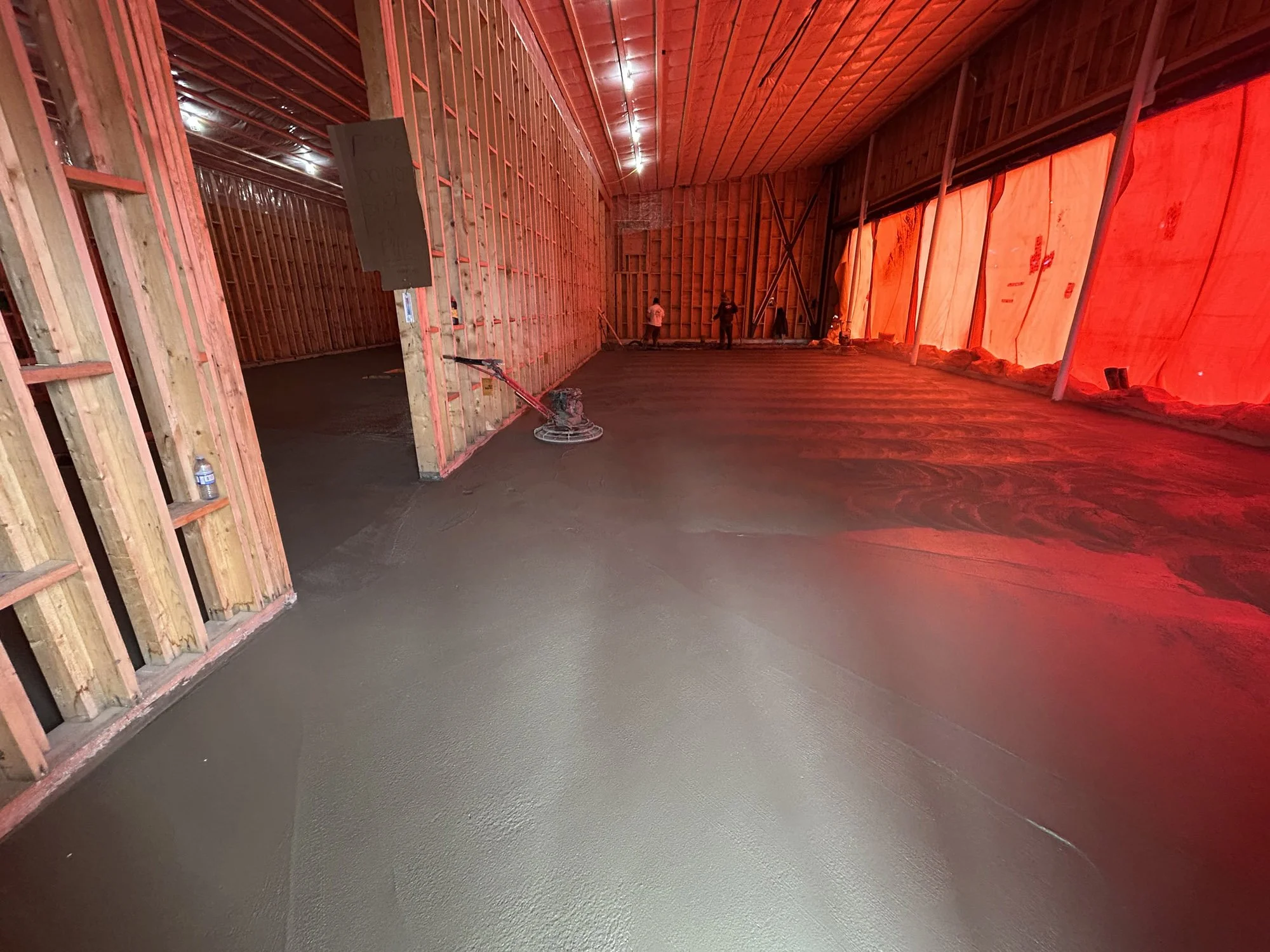 Interior of a building under construction with wooden framing and freshly poured concrete floor, illuminated by red lighting.