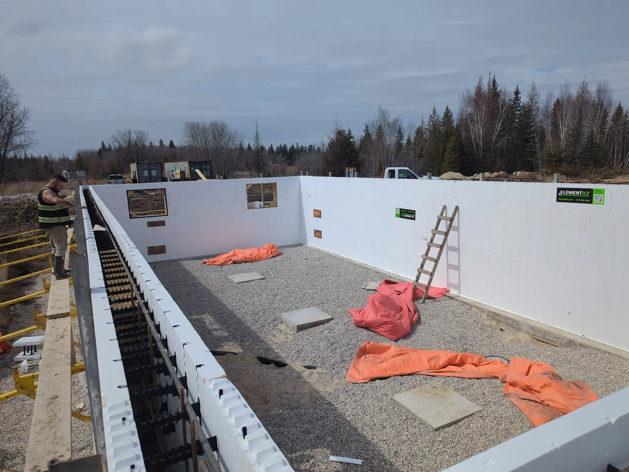 Construction site of a building with insulated walls, a ladder, and construction materials, including orange tarps and gravel ground, with a worker on the left side wearing a yellow safety vest and work boots.