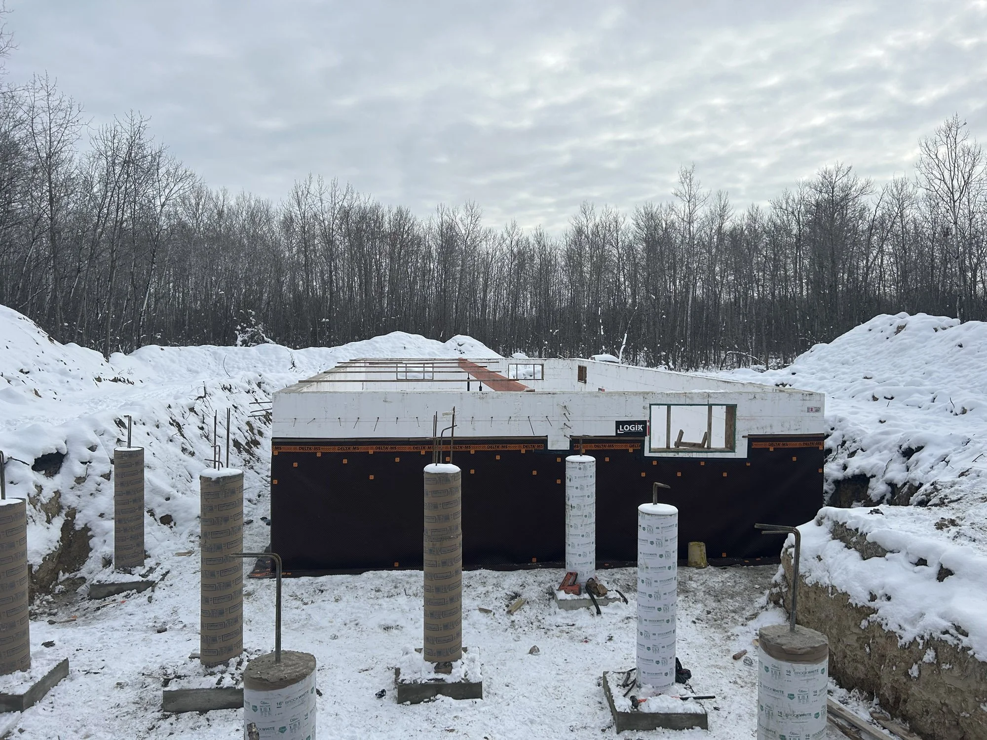 Construction site in winter with snow, showing a partially built structure with insulation material and metal framing, surrounded by snow-covered ground and trees in the background.