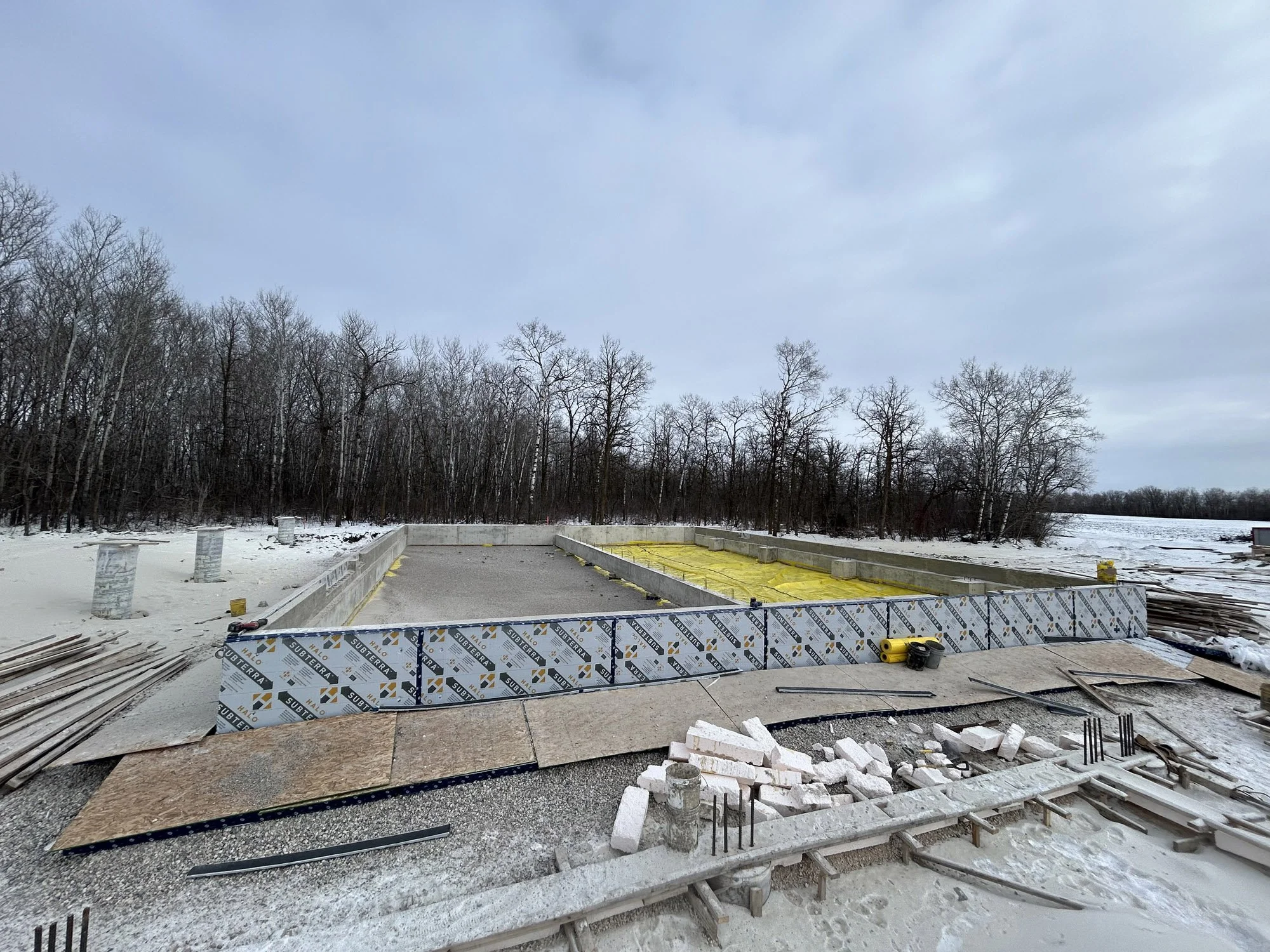 Construction site with foundation pouring in a snowy landscape, surrounded by leafless trees and cloudy sky.