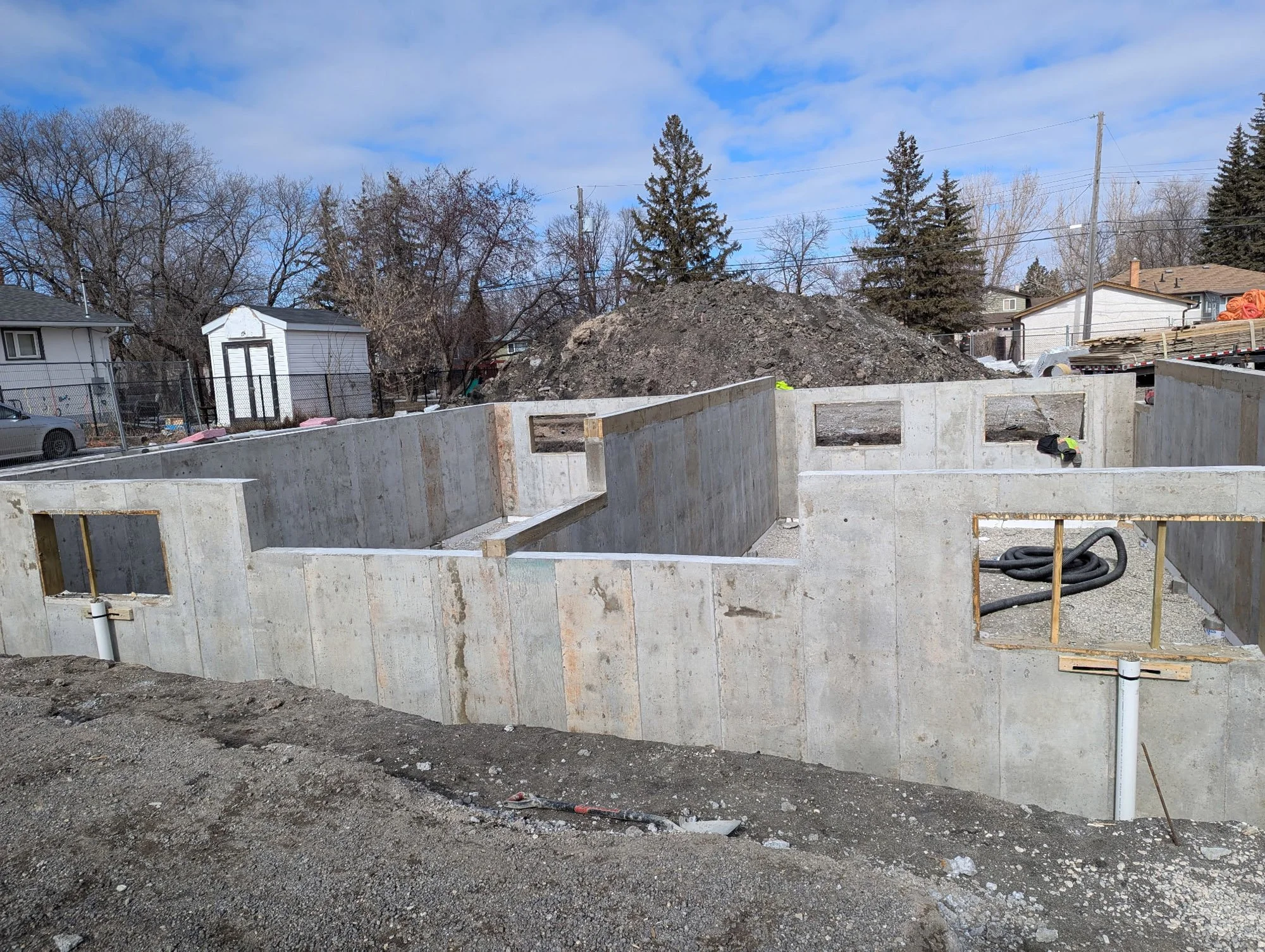 Construction site with concrete foundation walls for a building, with window openings, black hoses, and a small hill of dirt in the background, under a partly cloudy sky.