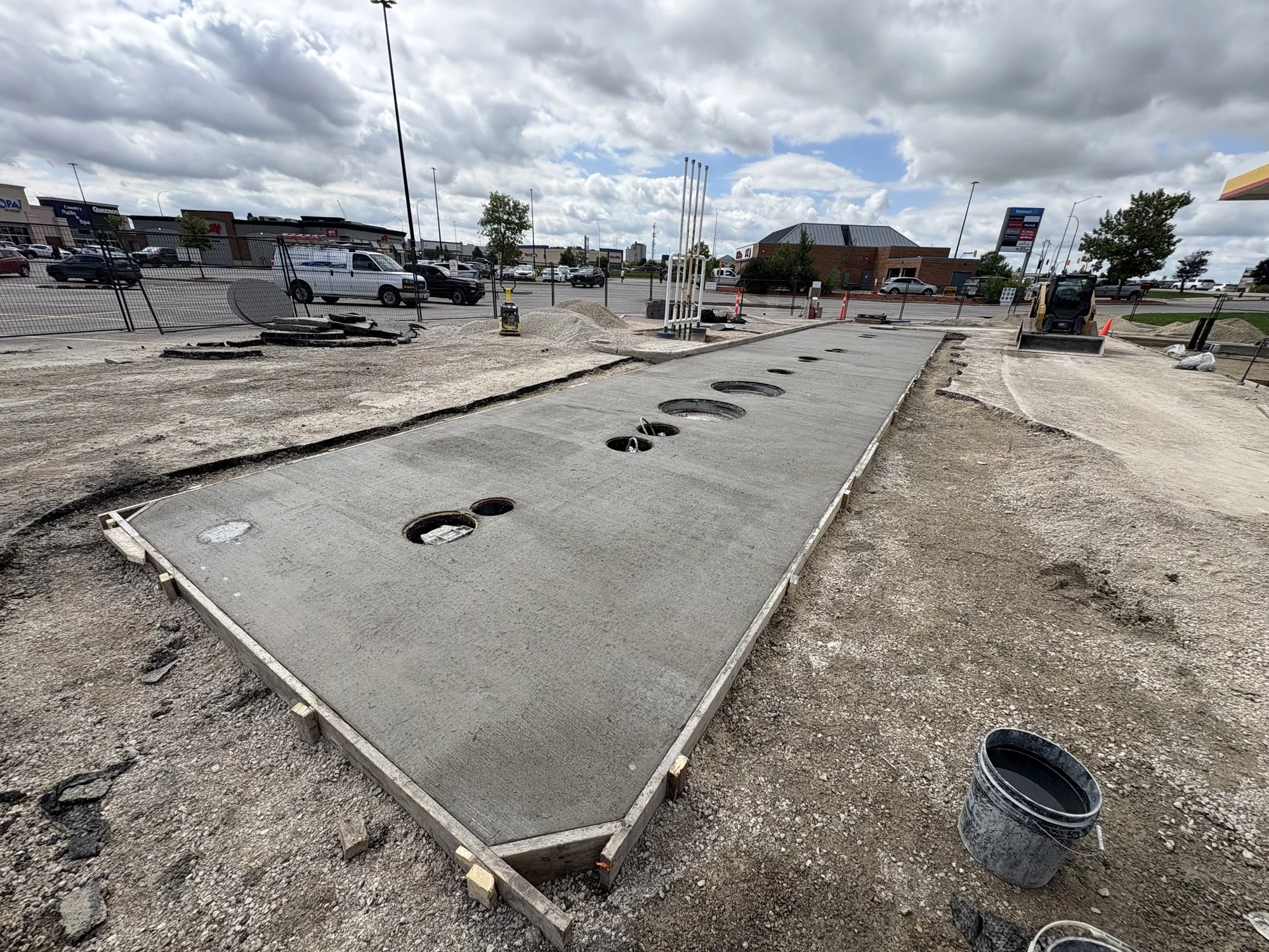 Construction site with freshly poured concrete sidewalk, circular manhole covers, construction equipment, and surrounding dirt, with parking lot and buildings in the background under cloudy sky.