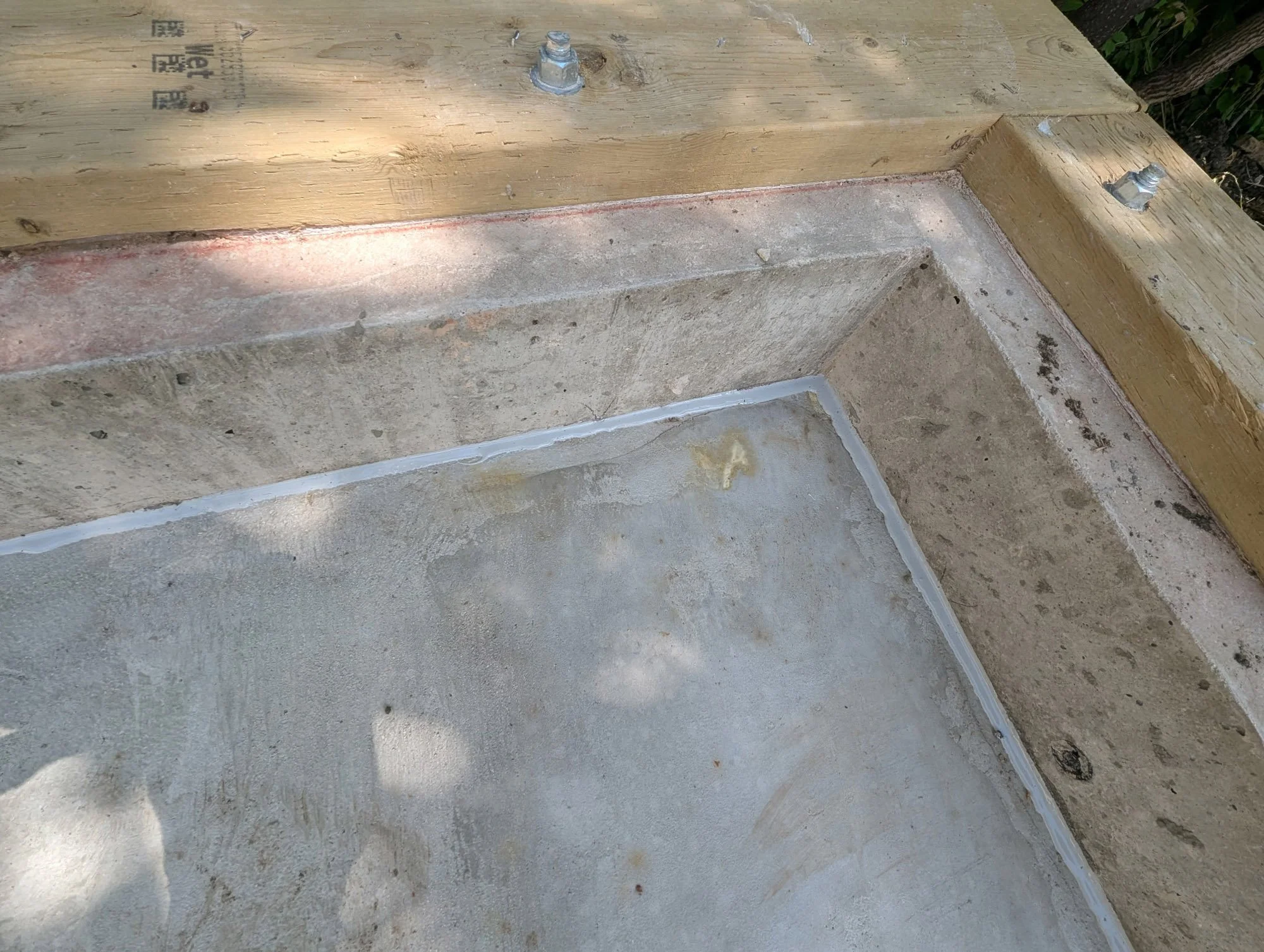 Close-up of a corner of a concrete structure with a visible joint sealed with white waterproofing material, framed by a wooden beam secured with bolts.
