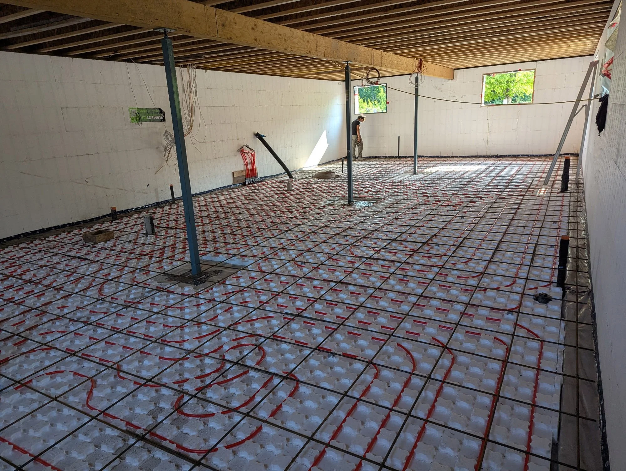 Interior of a building under construction with radiant floor heating pipes, metal support beams, and a worker inspecting the space near open windows.