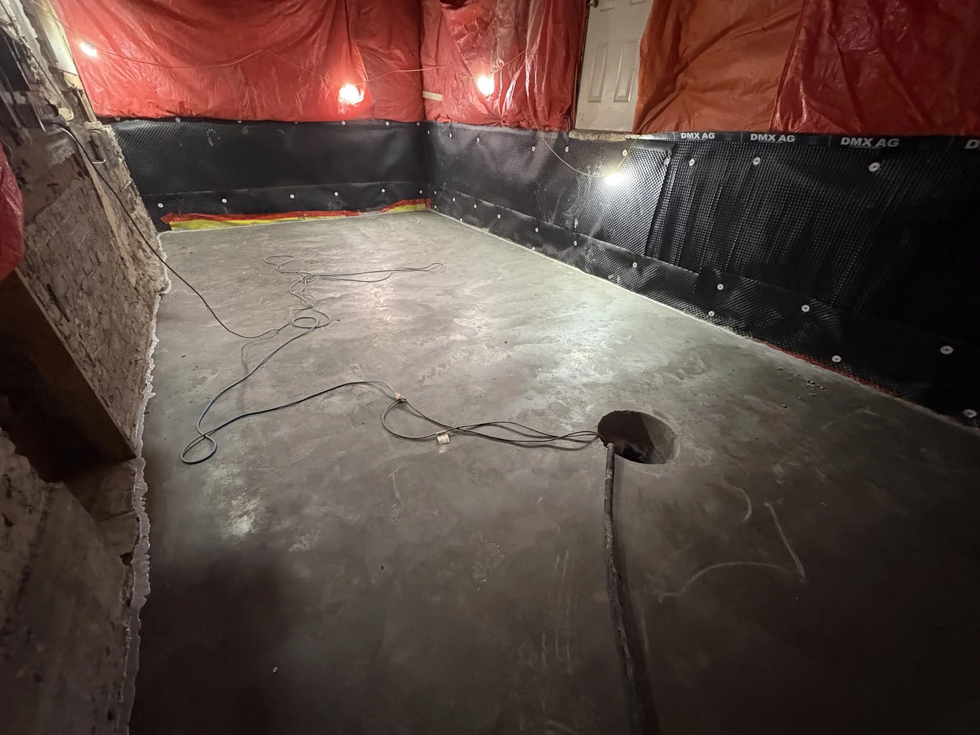 Empty basement with a hole in the concrete floor, electrical cords, and black waterproofing material on the walls. Red tarp draped on the walls with lights attached.