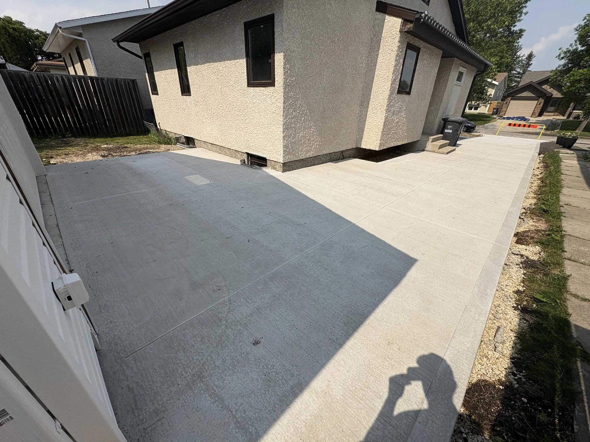Newly paved concrete driveway leading to the back of a beige house with black window frames. The driveway is partly shaded, and there is a shadow of the person taking the picture. A black fence and a grassy area are visible on the left, with some ste