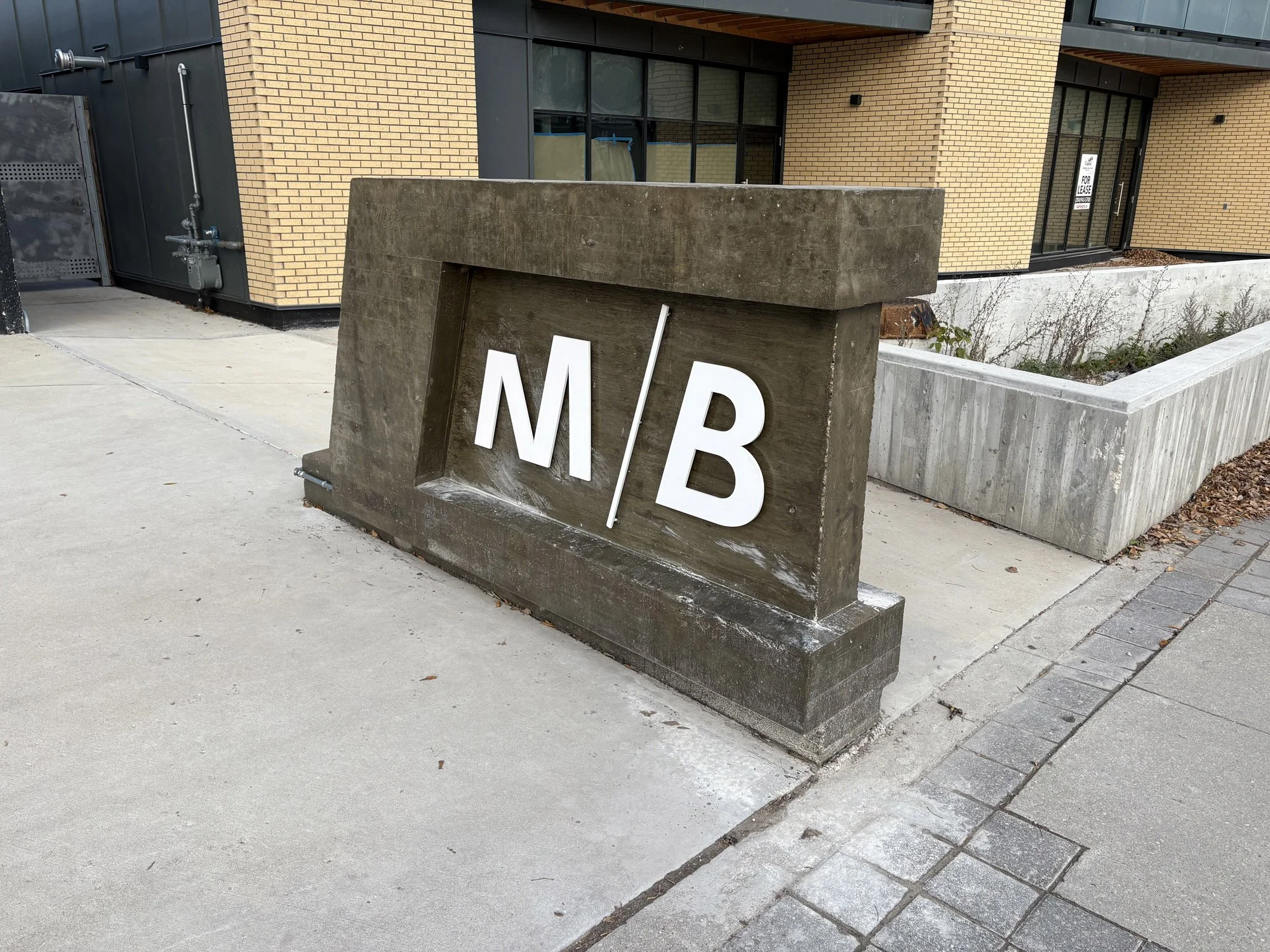 Concrete sign with large white letters 'M / B' engraved, situated on a sidewalk outside modern building.