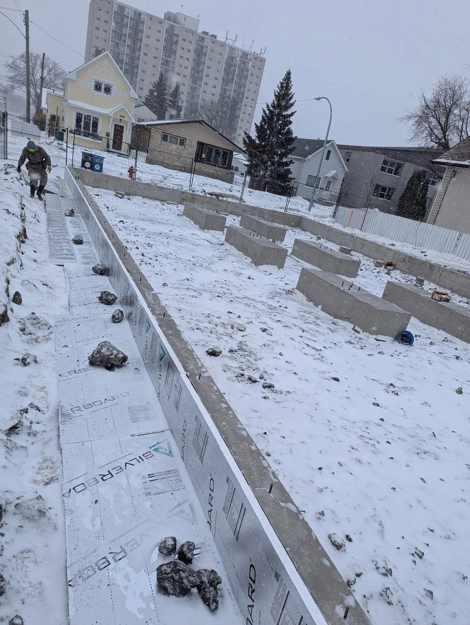 Construction site with concrete footings on snow-covered ground, a worker in winter gear, and residential houses and apartment buildings in the background during snowfall.