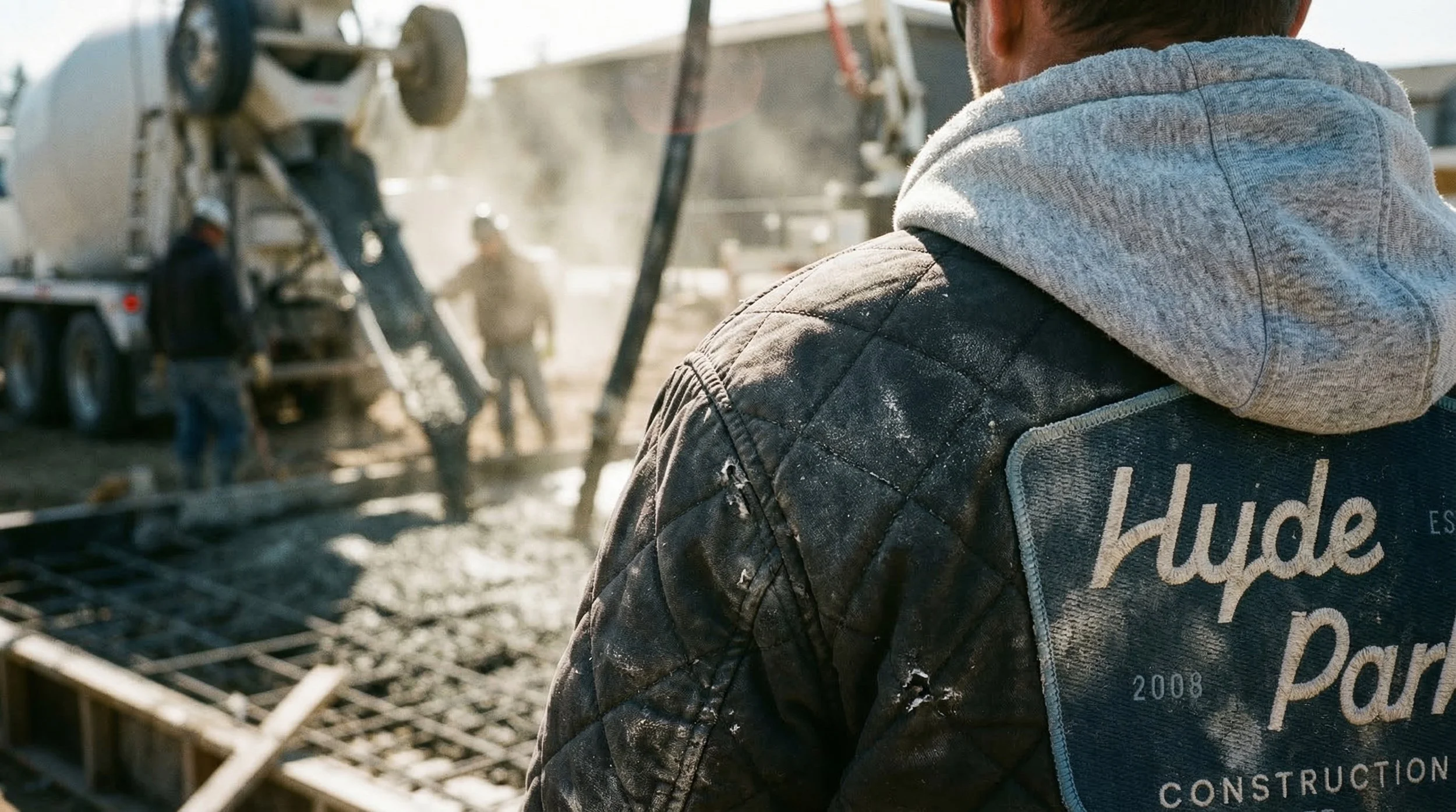 Close-up of a worker wearing a "Hyde Park" patched jacket and hoodie, observing construction workers pouring concrete at a construction site, with a cement mixer truck in the background.