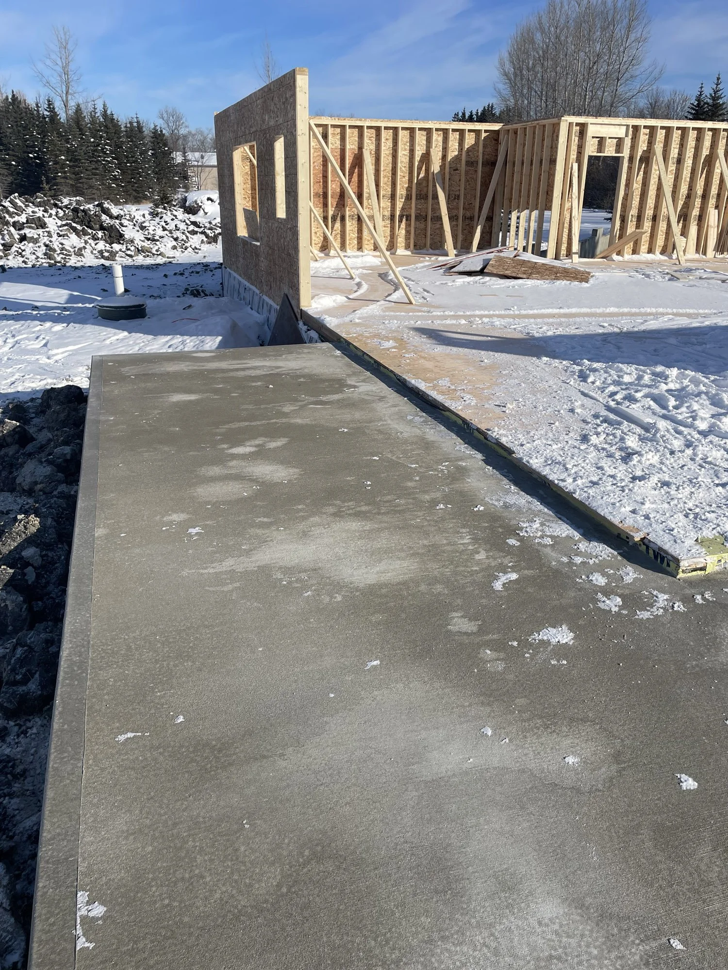Construction site in winter with partially built wooden framing of a house, concrete foundation, and snow-covered ground.