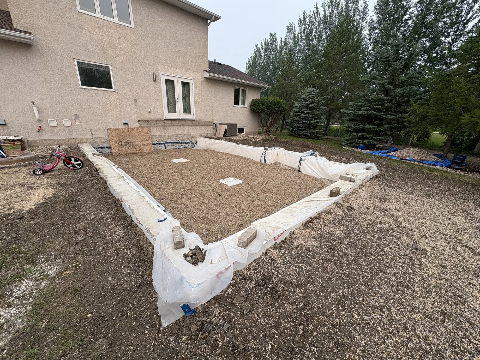 A residential backyard undergoing construction with a framed foundation, gravel, bricks, and construction materials visible. A pink children's tricycle is on the left side, and blue tarps cover some materials in the background. The house has beige st