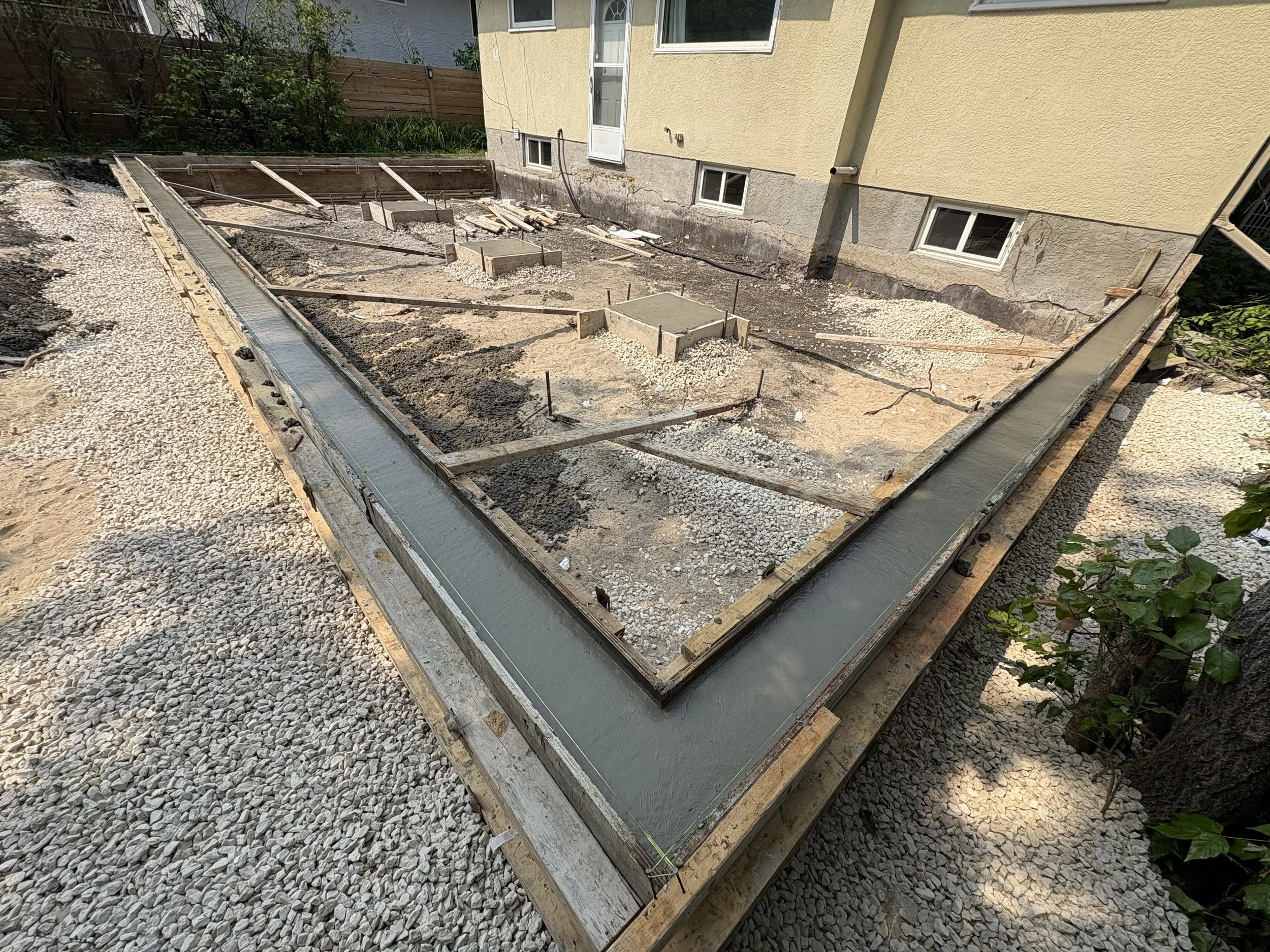 Construction site showing the foundation framework with fresh concrete in a residential backyard, surrounded by gravel and dirt, next to a yellow house.