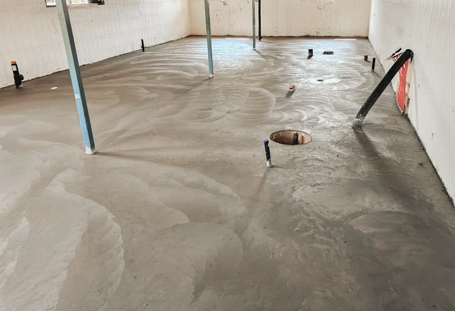 Interior of a room under construction with a finished cement floor, plumbing pipes protruding from the floor, and support beams, with walls ready for finishing.
