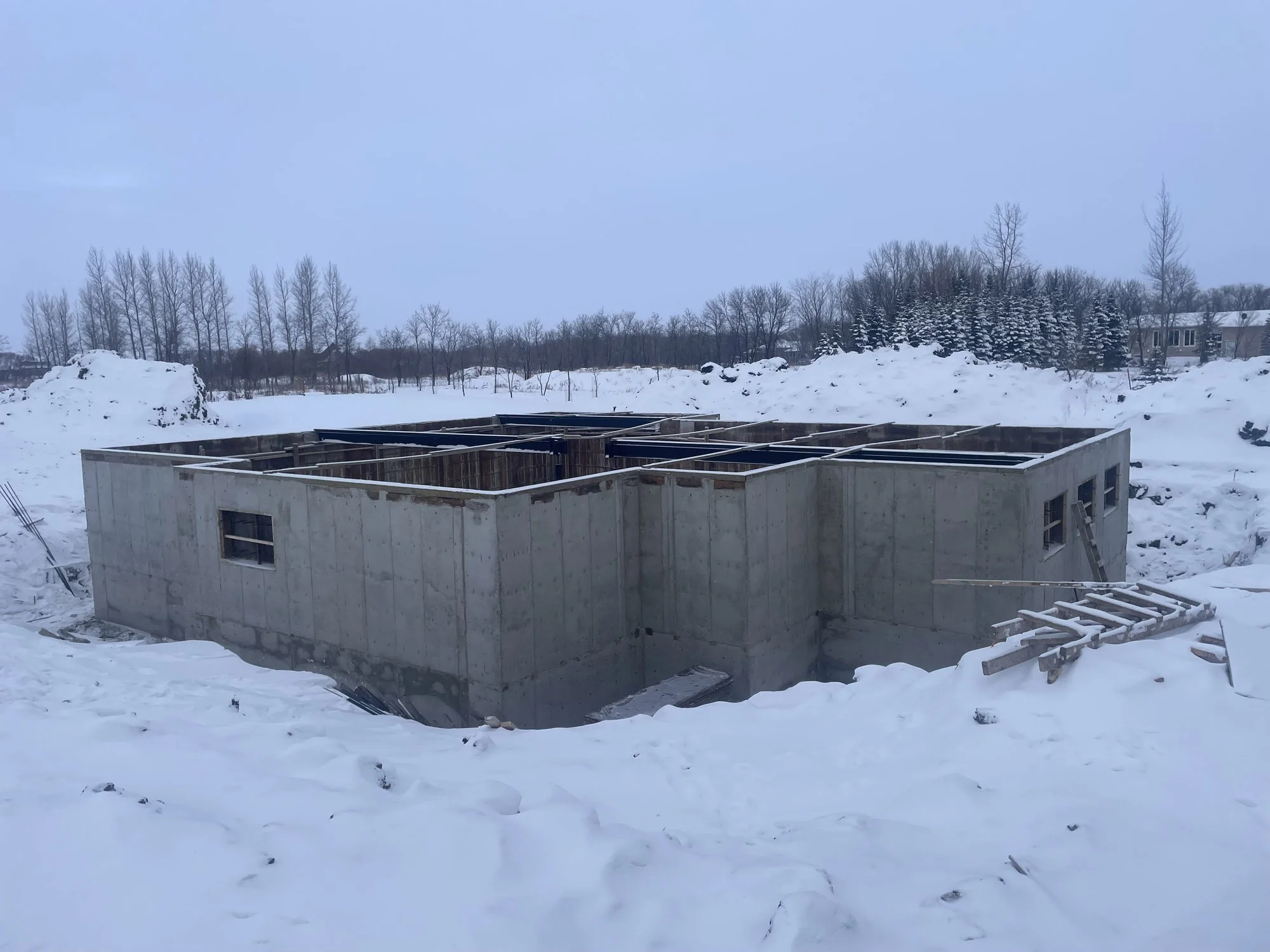 Snow-covered construction site with a concrete foundation outline for a building, surrounded by snow and trees in the background during winter.